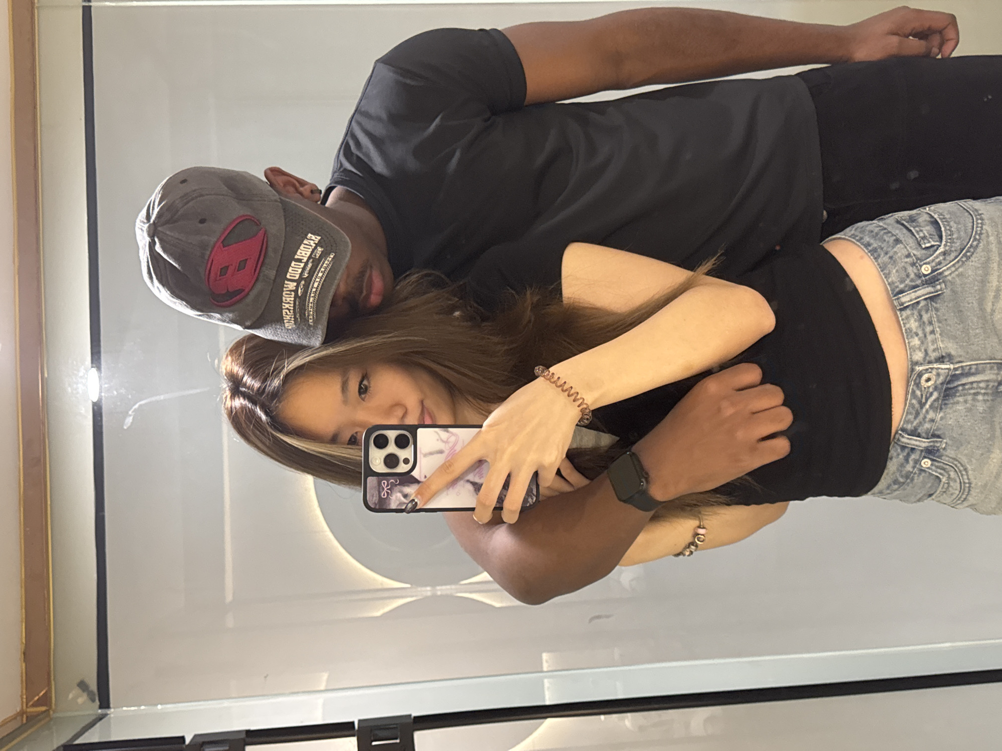 A selfie of a young couple is reflected in a mirror. The woman is holding a smartphone, capturing the image. She has long, light brown hair and is wearing a black top and light blue denim shorts. The man beside her wears a dark grey baseball cap with a red logo, a black t-shirt, and black pants. The background appears to be a bathroom or dressing room with white walls and a round light fixture. The location in Yuzhong District, China cannot be ascertained from the image alone.