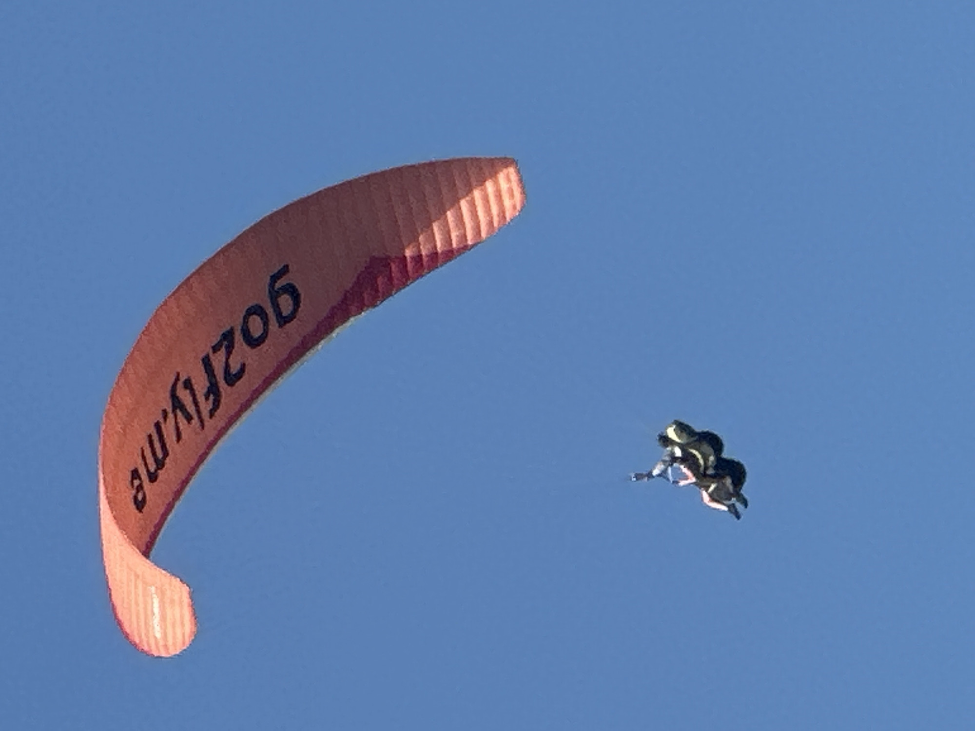 A paraglider with the logo "go2fly.me" is in mid-air against a clear blue sky. The paraglider is orange and has black lettering. Beneath the paraglider are two people, appearing to be tandem paragliding. They are suspended in harnesses, facing forward, with one person in front of the other. The background is a solid, cloudless blue sky, suggesting an outdoor activity in good weather. The image does not provide specific geographical information for Boreti, Montenegro.