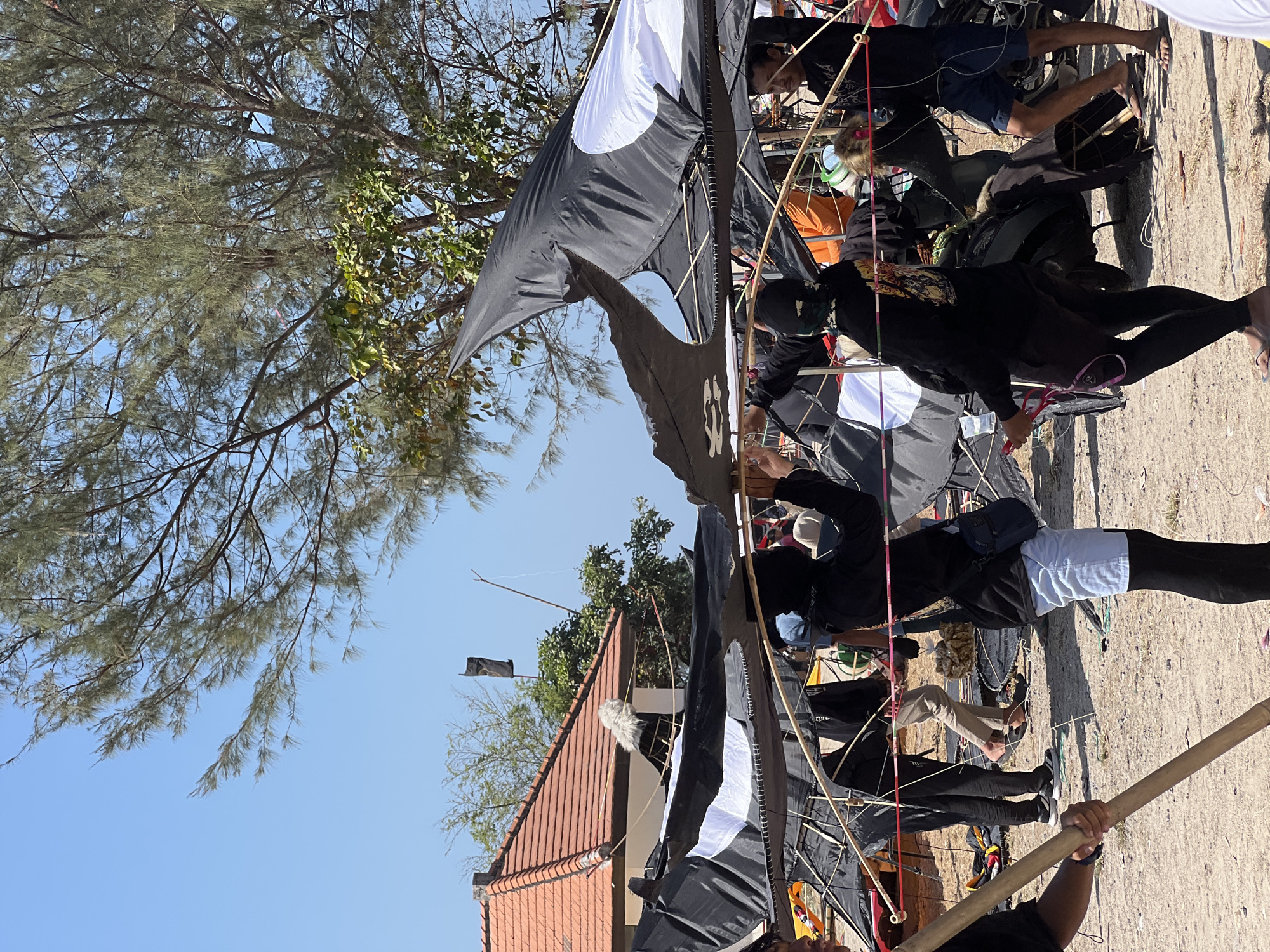 In Denpasar, Indonesia, a group of people are gathered on a sandy area near trees, preparing large black and white kites. The kites, some partially assembled and others appearing damaged, are made of fabric stretched over wooden frames. Several individuals, dressed casually in black shirts and shorts or light-colored pants, are seen holding parts of the kites or assisting in their construction. One person is holding a long wooden pole, while others are holding strings and fabric. In the background, a red-tiled roof building with white trim is visible, along with dense foliage from trees and a clear blue sky. The scene appears to be outdoors, possibly a beach or an open field, with evidence of kite-making activities.