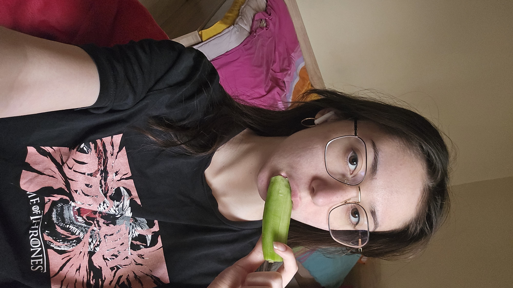 A woman is holding a green vegetable to her mouth. She is wearing a black t-shirt with a "Game of Thrones" graphic and gold-framed glasses. She has an earbud in her ear. The background shows a red blanket, a bed with colorful bedding, and a light-colored wall. There is no information to determine if the photo was taken in Stara Zagora, Bulgaria.
