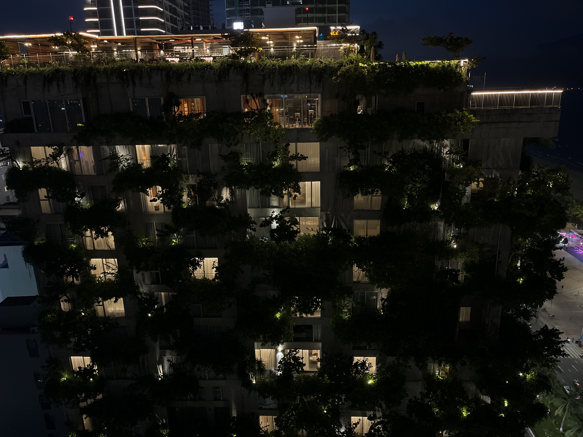 At night, the image shows a tall, concrete building covered in greenery in Đà Nẵng, Vietnam. The building has multiple stories, with windows lit from within. Some windows reveal activity, suggesting occupancy. Above the building is a rooftop terrace with lights and greenery. In the background, taller buildings are visible, as is a glimpse of the coastline and city streets. The streets show lights and vehicular activity. The overall ambiance is urban and tropical, with a modern building integrated with nature.
