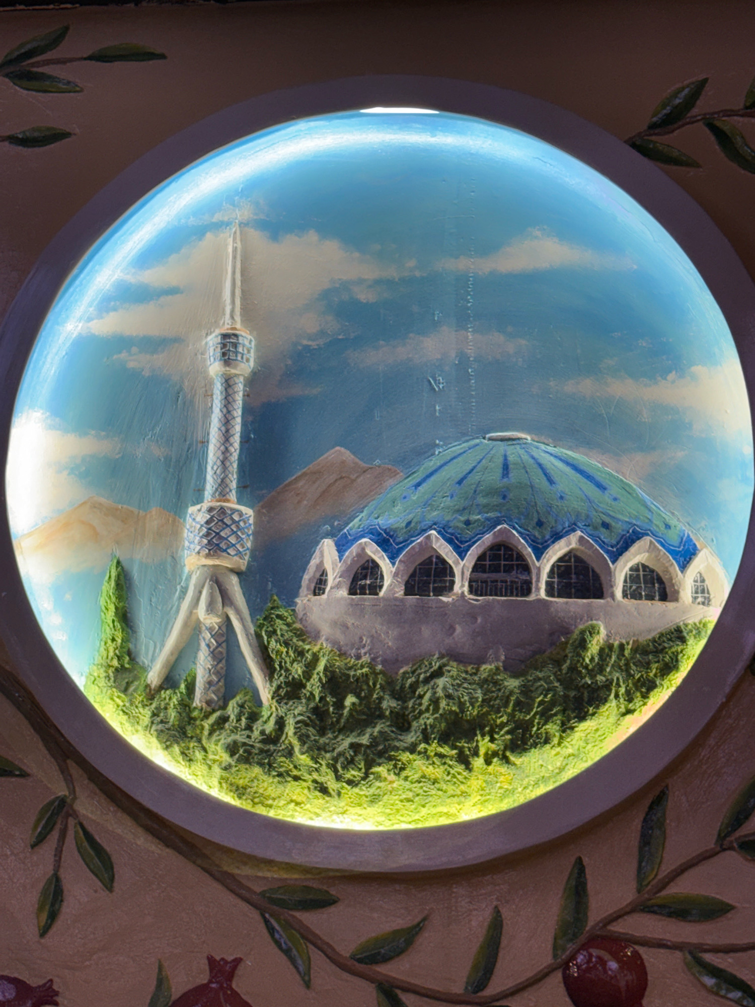 The image shows a circular bas-relief artwork depicting the Tashkent Television Tower and the Alisher Navoi Opera and Ballet Theatre against a backdrop of mountains and sky. The Television Tower, a prominent landmark in Tashkent, Uzbekistan, is shown with its lattice structure and antenna. To the right, the dome of the Alisher Navoi Opera and Ballet Theatre is visible, with arched windows along its base. The foreground of the artwork features a textured, green landscape with trees. The artwork is framed by decorative branches with leaves and pomegranates, suggesting it is part of a larger mural or decorative element. The lighting within the circular artwork highlights the three-dimensional aspects of the relief.