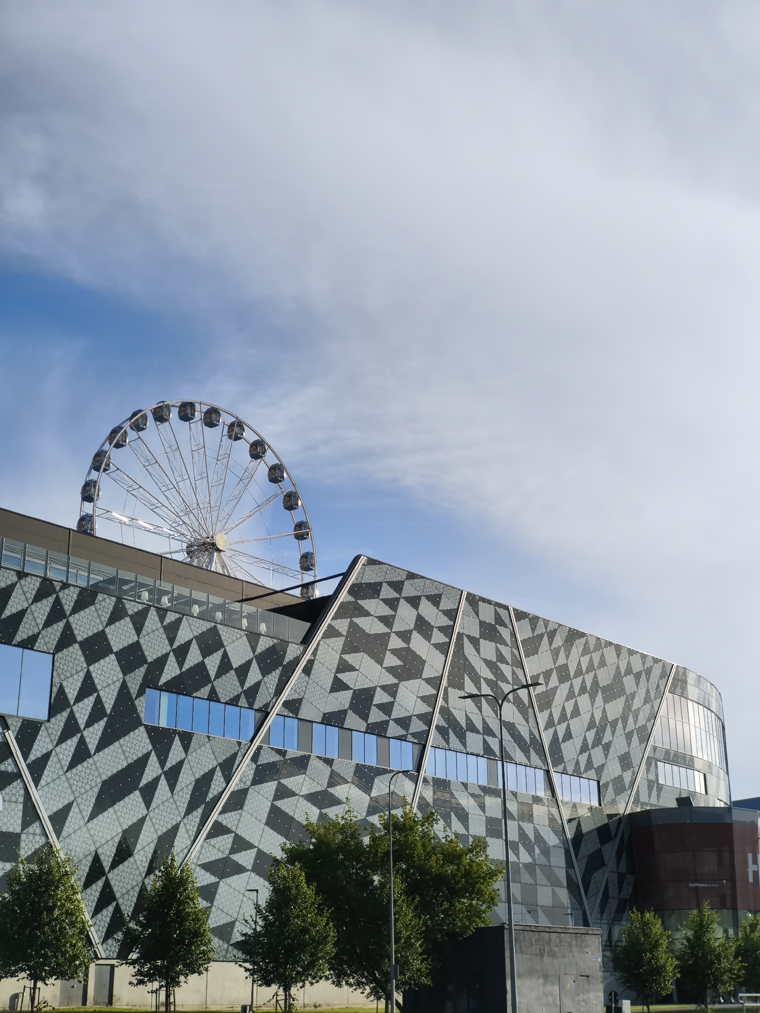 The image shows a modern building with a geometric facade, featuring a pattern of dark and light grey triangles. The building has large windows, some of which reflect the blue sky and clouds. A Ferris wheel, appearing to be silver and white, is visible in the background, peeking over the top of the building. Several trees with green foliage line the front of the building. The sky is blue with scattered white clouds, with a large portion of the image dedicated to the sky. The scene appears to be an outdoor public space in the city of Tallinn, Estonia, possibly near a commercial or entertainment complex.