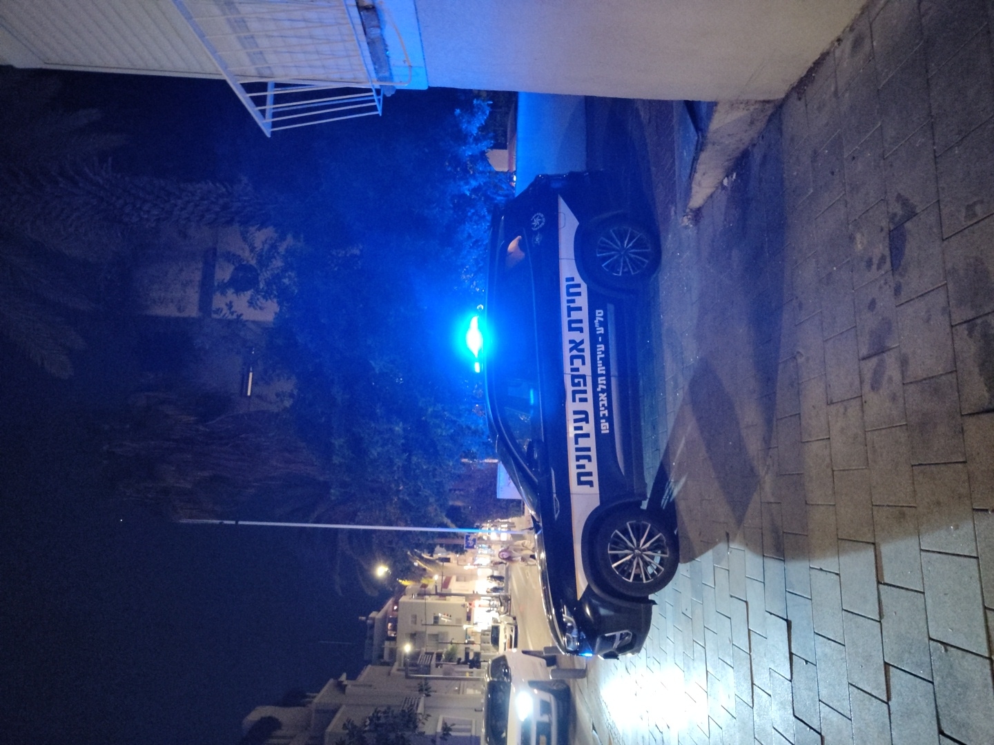 A dark-colored municipal enforcement SUV is depicted at night on an urban street in Tel Aviv-Yafo, Israel. The vehicle displays Hebrew text along its side, reading "יחידת אכיפה עירונית" (Municipal Enforcement Unit) and "סל"ע - עיריית תל אביב יפו" (Sela - Municipality of Tel Aviv-Yafo). A blue emergency light on the vehicle's roof is active, casting a vivid blue illumination onto the vehicle and surrounding environment. The SUV is stopped on a paved sidewalk composed of grey square tiles, adjacent to the street. In the background, multi-story buildings with illuminated windows line the street, where streetlights are also visible. A white vehicle is partially visible on the far left of the street. Indistinct human figures are present further down the street to the left. Dense foliage, including palm trees, is visible behind the enforcement vehicle, partially obscured by the darkness and the blue light. A building section with a white-railed balcony is visible on the far right.