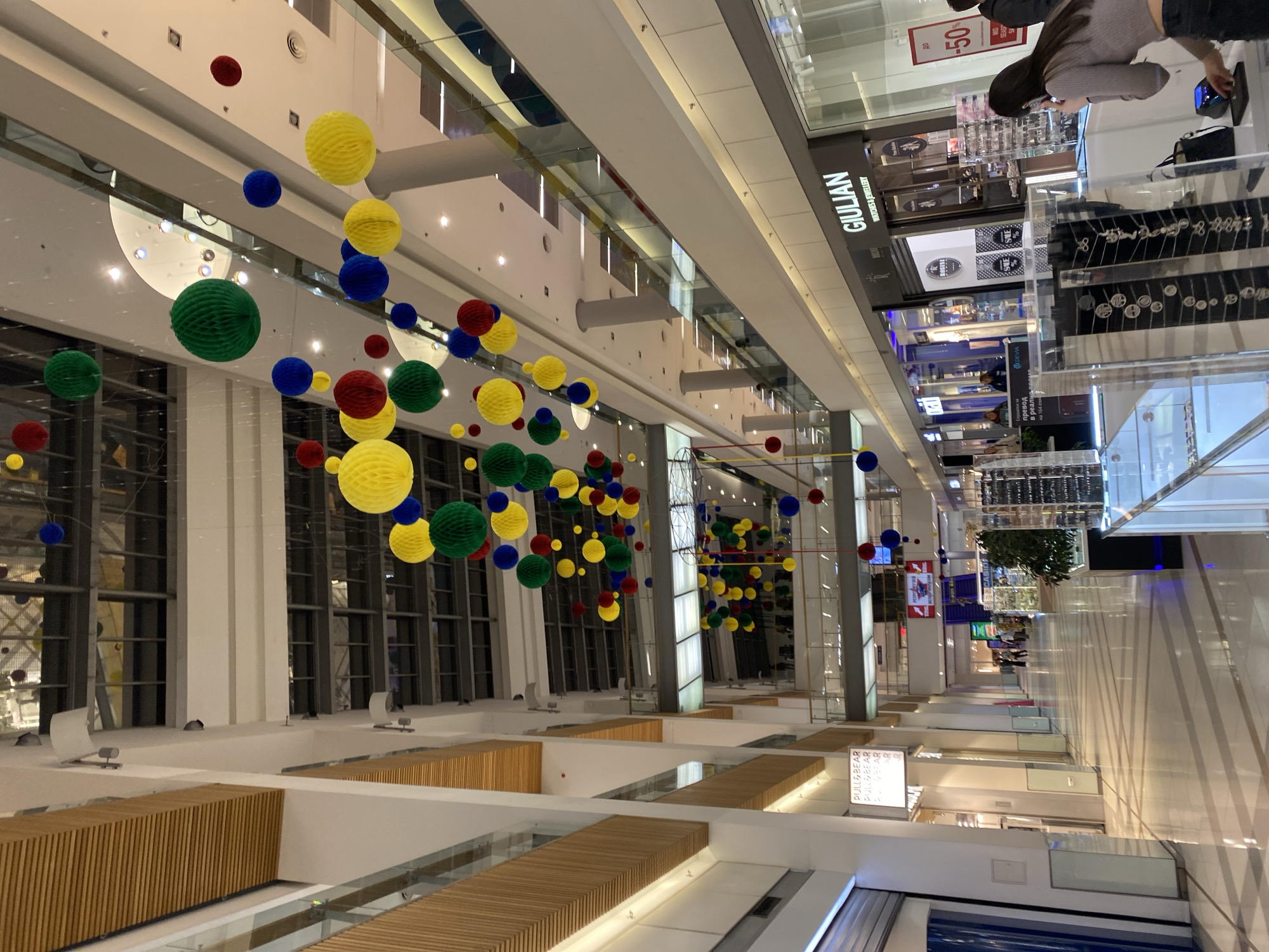 This is an interior view of a bright, multi-story shopping mall or large commercial complex in Varna, Bulgaria. The image captures a wide-angle perspective, looking upwards and along a spacious corridor or atrium.
The most prominent feature is the vibrant decoration: numerous honeycomb paper balls or lanterns in red, yellow, green, and blue are suspended at various heights from the high ceiling. These colorful spheres create a festive and lively atmosphere throughout the space.
The mall features modern architecture with clean, white walls, large glass railings for balustrades on upper levels, and wooden slatted accents along some ledges. Natural light seems to enter from upper windows or skylights, complemented by extensive artificial lighting from recessed fixtures.
On an upper level, a person, likely a woman with long dark hair, wearing a gray top and dark pants, is visible looking into a glass display case, possibly browsing merchandise. Further down the corridor, other indistinct figures can be seen, indicating ongoing activity within the mall.
Several store signs are visible. On one upper level, a prominent black sign reads "GIULIAN WATCHES & JEWELLERY." Nearby, a red sign advertises "до -50%" (up to -50% off), indicating a sale. Another store sign for "PULL&BEAR" is visible on a lower level. Other Cyrillic text, "превод в реаль на 164," is also present, likely an advertisement or service offering. The scene appears well-maintained and orderly.