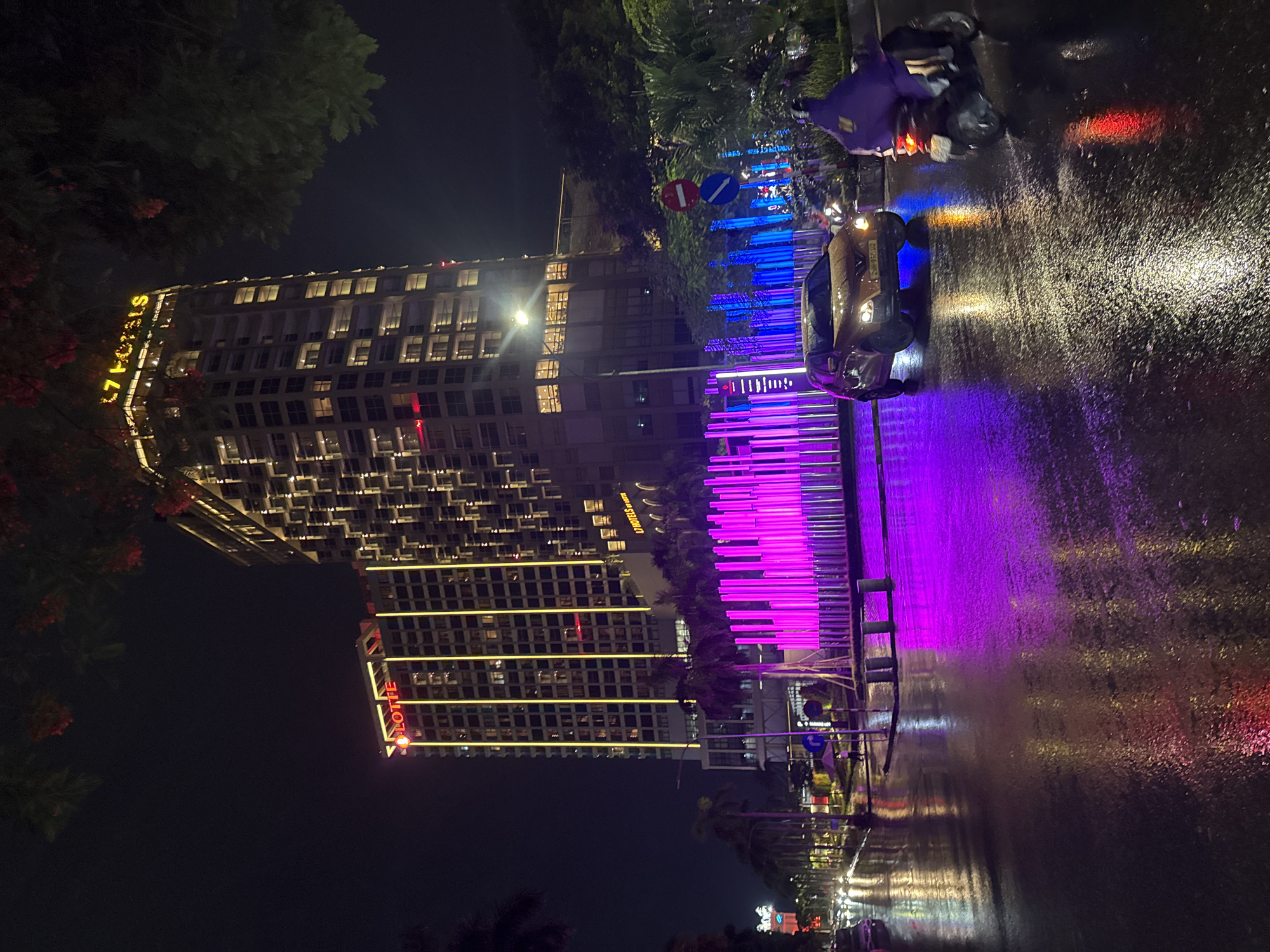 The image depicts a nighttime street scene in Hanoi, Vietnam, with a focus on the Lotte Hotel. The Lotte Hotel, a tall, multi-story building, is illuminated with vertical yellow lights and displays a prominent "LOTTE HOTELS" sign in red and yellow lettering at its peak. The street in front of the hotel is wet and reflective, indicating recent rainfall. A golden-colored car is driving on the wet street, its headlights on. To the right, a person wearing a purple rain jacket is riding a scooter, also on the wet road. In the foreground, a vibrant display of purple and blue lights emanates from an installation, casting reflections on the wet asphalt. Palm trees are visible along the street, and distant city lights and traffic can be seen in the background. The overall atmosphere is one of a bustling city night with wet conditions.
