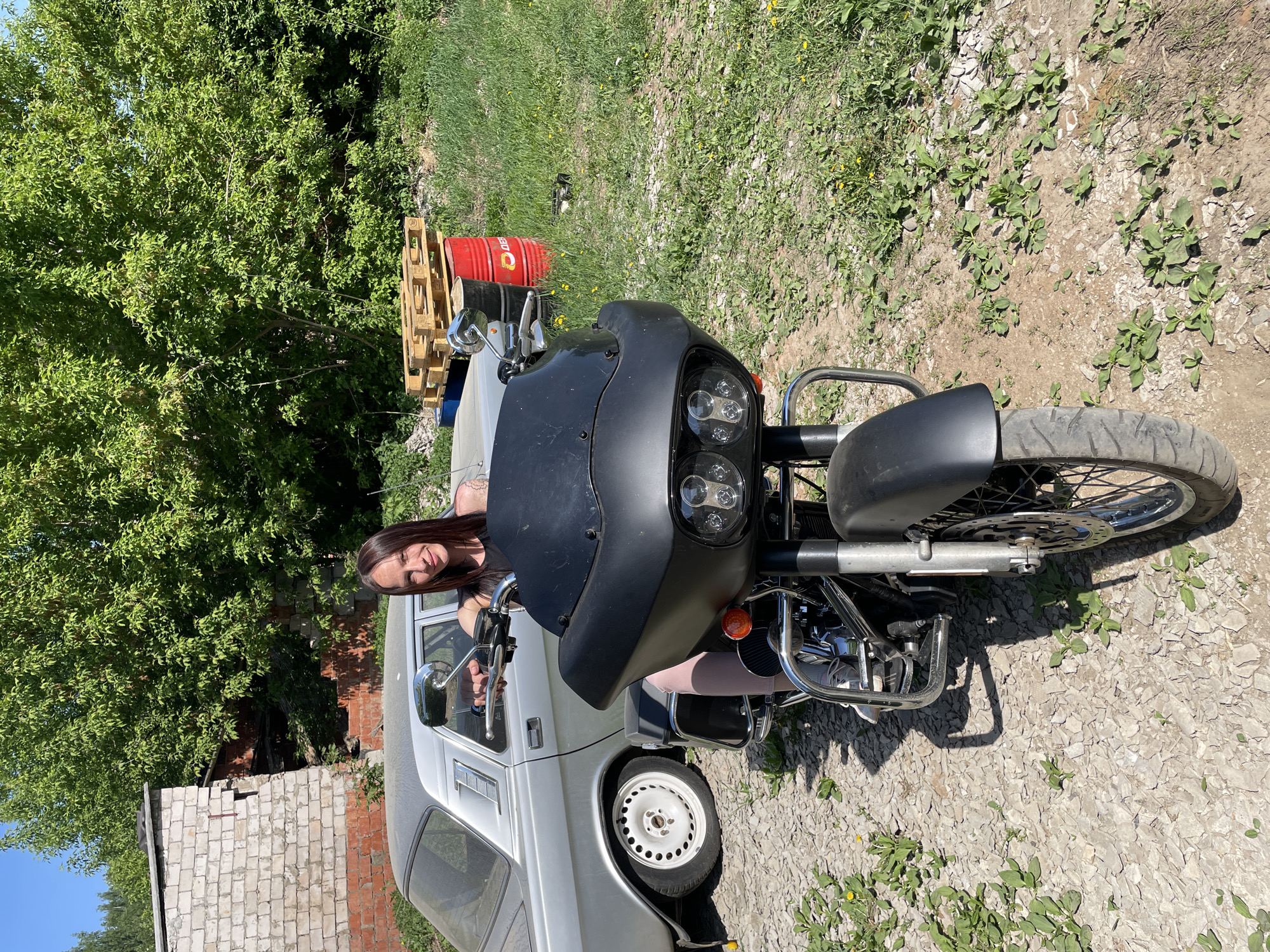A young woman with long dark hair is seated inside a silver car with the driver's side window rolled down. A black motorcycle is parked directly in front of the car, obscuring part of the vehicle. The scene is set outdoors, with a ground cover of gravel and sparse vegetation. Behind the car, to the left, there is a small brick building. Further back, a lush green tree is visible. To the upper left of the car, wooden pallets and a red metal barrel are situated. The location is identified as Oktyabr'skii, Russia.