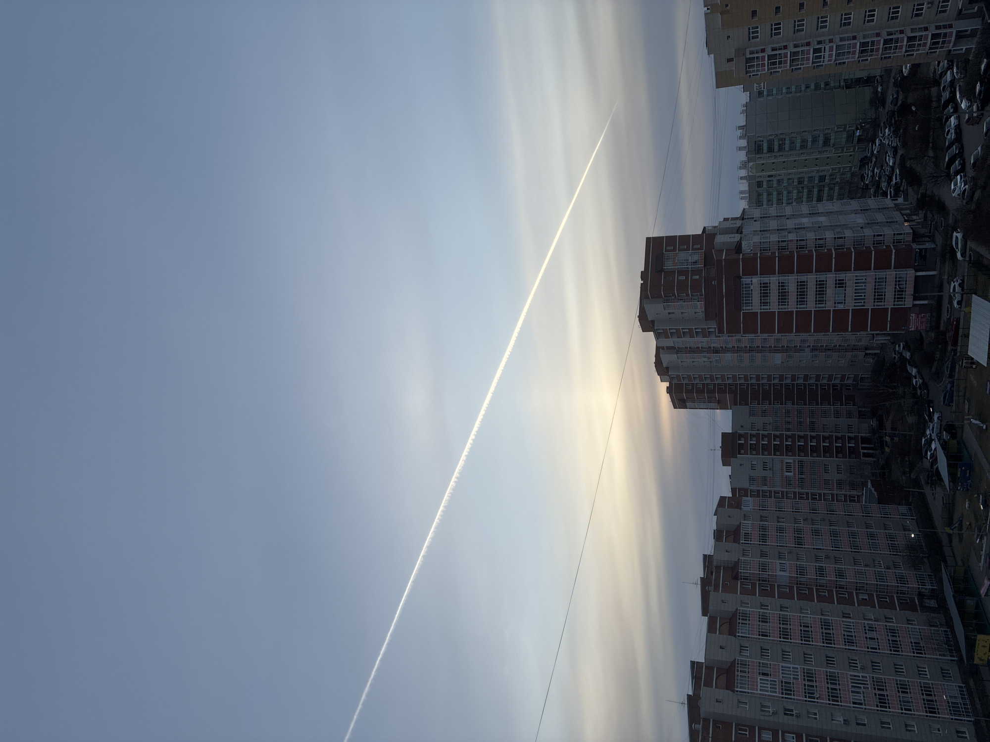 The image depicts a cityscape under a partly cloudy sky with a visible contrail from an airplane. Several tall, multi-story apartment buildings dominate the right side of the image, with a visible parking area and cars below. Power lines are faintly visible across the sky. The buildings are characterized by their uniform windows and red and gray color schemes. The overall scene appears to be taken during the day, with adequate sunlight illuminating the buildings.
