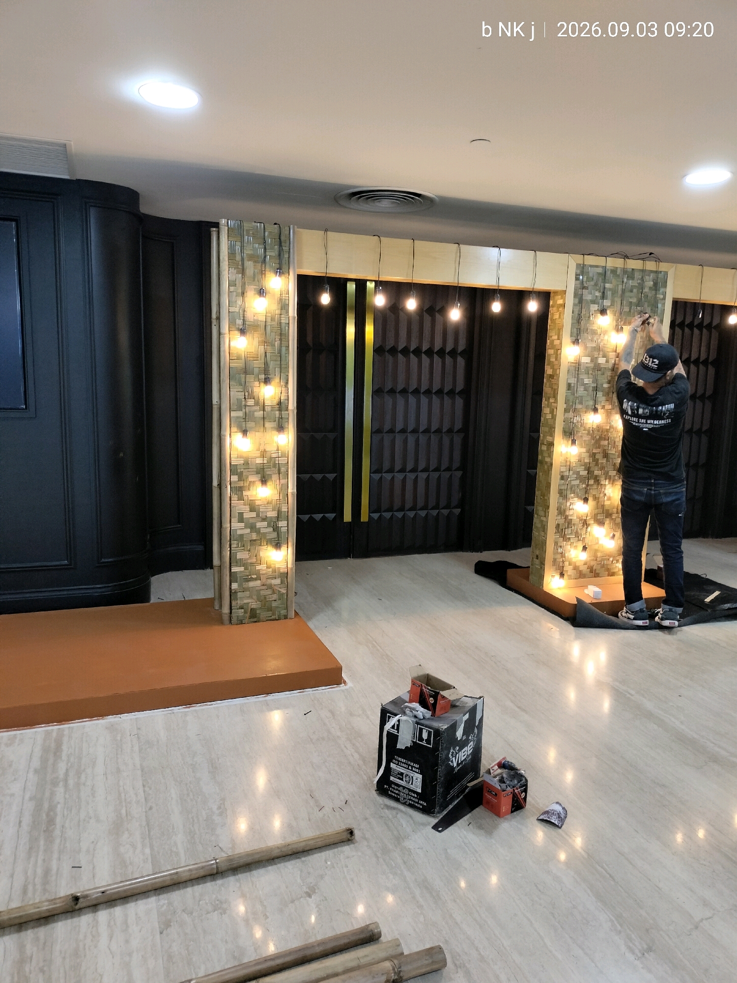 The image depicts an indoor scene, likely a construction or renovation site in South Jakarta, Indonesia, with the timestamp indicating September 3, 2026, at 09:20. A person, dressed in a black t-shirt and jeans, is actively engaged in attaching string lights to a decorative wall structure. The wall itself is characterized by a woven bamboo-like material, interspersed with numerous illuminated light bulbs, creating a festive atmosphere.
To the left, a dark, paneled wall with a curved edge suggests a sophisticated interior design element. In front of this, a raised platform with an orange surface hints at a stage or display area. The floor is a polished marble with a visible pattern. Scattered on the floor are construction materials, including cardboard boxes, tools, and what appear to be bamboo poles, indicating ongoing work. The overall environment suggests an event preparation or interior finishing project.