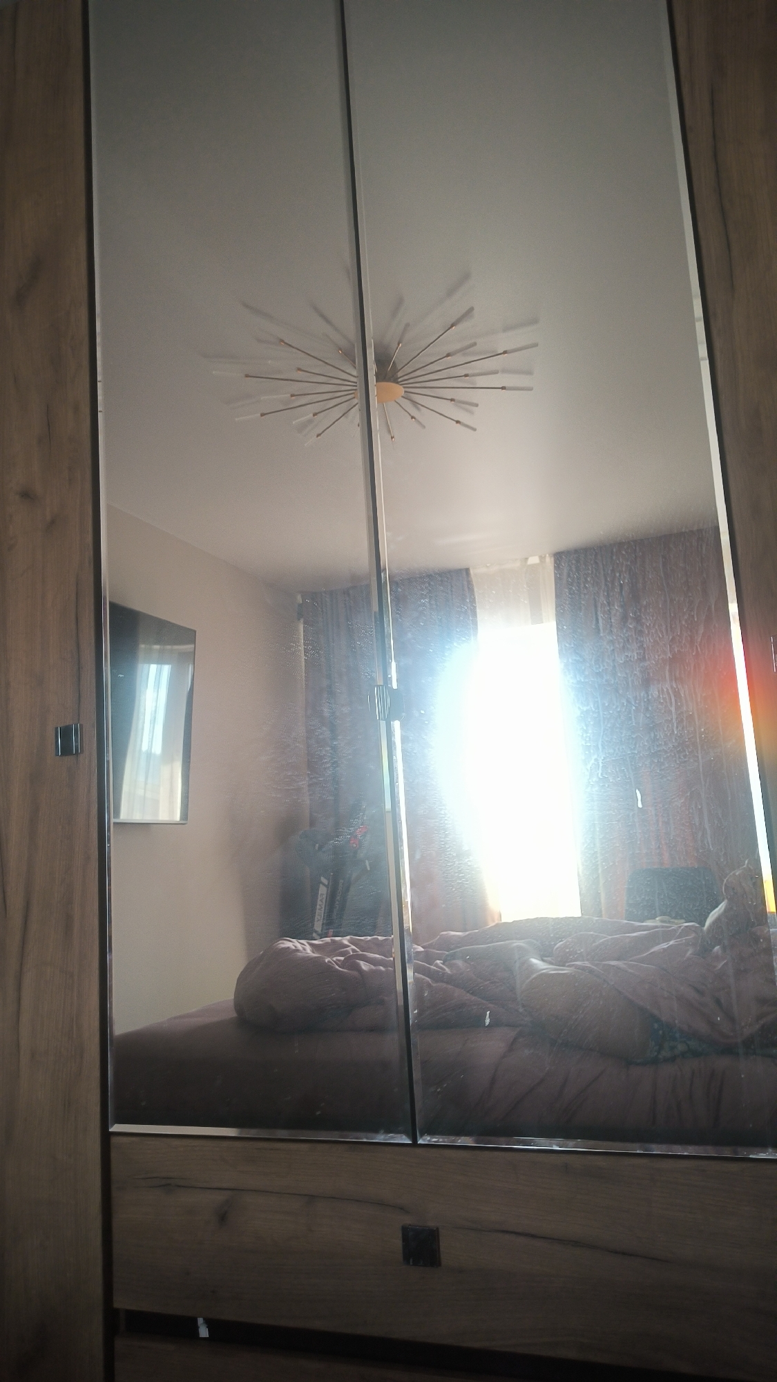 This is a vertical, medium-shot, eye-level photograph of a bedroom reflected in a large mirrored wardrobe. The wardrobe has a wooden frame and two mirrored doors. The reflection shows a bed with rumpled pink bedding, a window with brown curtains, and a ceiling light fixture that resembles a starburst. There is a television mounted on the wall to the left. Sunlight streams in through the window, creating a bright glare that obscures some details. The room appears to be lived in, with clothes draped over furniture. The overall impression is of a casual, domestic setting. The time of day appears to be morning or afternoon, given the strong sunlight. There is no visible text in the image. The location context is Kovrov, Russia.