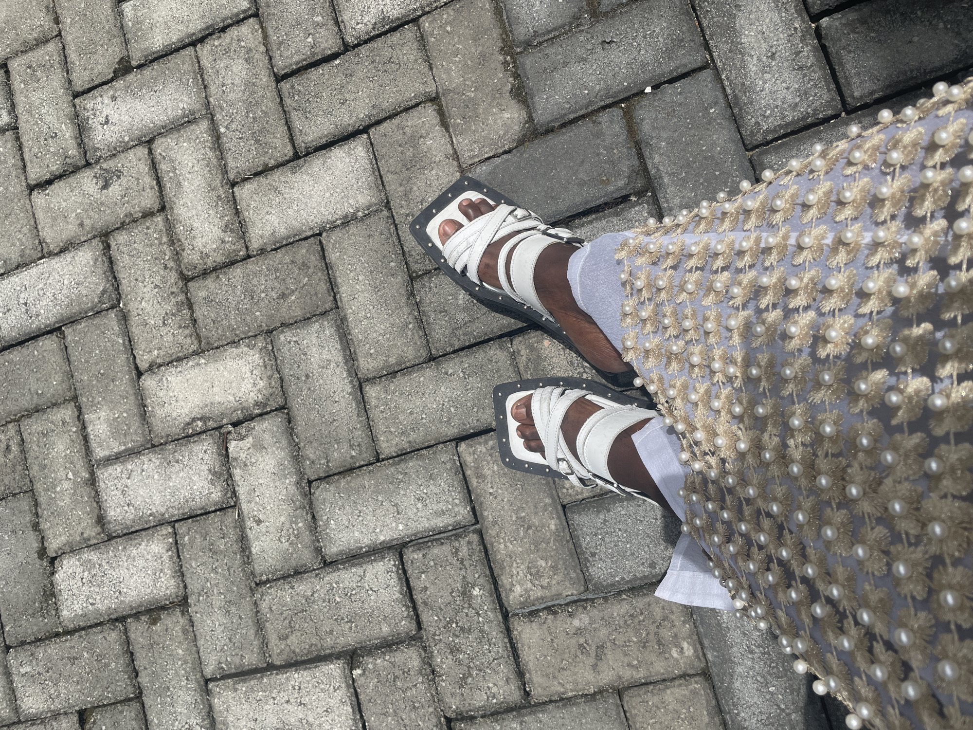 The image shows a person standing on a paved area, likely a sidewalk or pedestrian walkway, constructed of interlocking grey brick pavers arranged in a herringbone pattern. The person is wearing white sandals with a square toe and multiple straps, and is wearing what appears to be a pale blue or white garment embellished with small, evenly-spaced pearls and floral embroidery. The location is possibly in Lekki, Nigeria.
