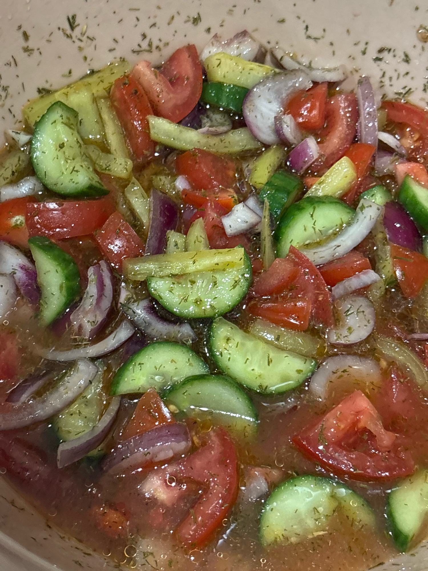 The image shows a close-up view of a salad in a light-colored bowl. The salad appears to be composed of chopped tomatoes, cucumbers, and red onions, all tossed in a dressing. Dried herbs, likely dill, are visible throughout the salad. The lighting is overhead, creating reflections on the surface of the dressing. The salad is in a bowl, which is partially visible at the bottom and edges of the frame. There are no people or animals present. The setting does not provide any specific information about the city of Yuzhny, Russia, or any urban environment.