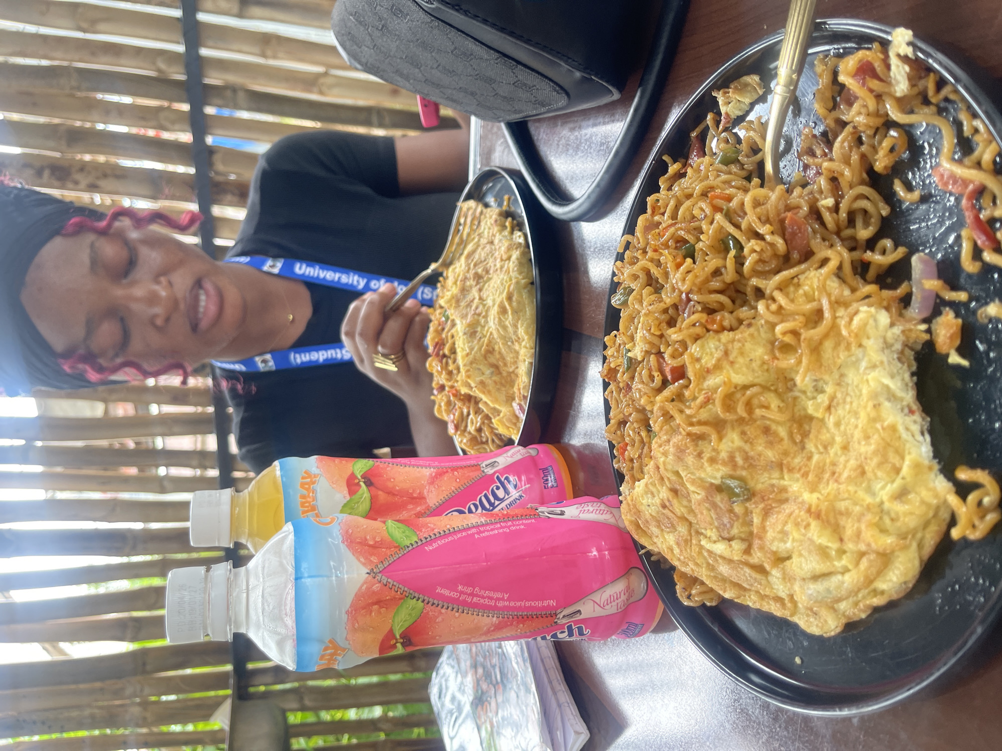 This image depicts a close-up view of a meal on a table, with a person in the background. The table is set with two black plates containing food, a bottle of peach-flavored juice, and a purse. The food on the plates appears to be instant noodles mixed with vegetables and topped with an omelet. The person in the background is wearing a black shirt and a headscarf, and has pink curly hair. They are looking down at their food, with a fork in their hand. The environment appears to be an outdoor setting with a bamboo fence in the background, suggesting a casual dining area. The overall scene suggests a person enjoying a meal. The location is identified as Rafin Par, Nigeria.