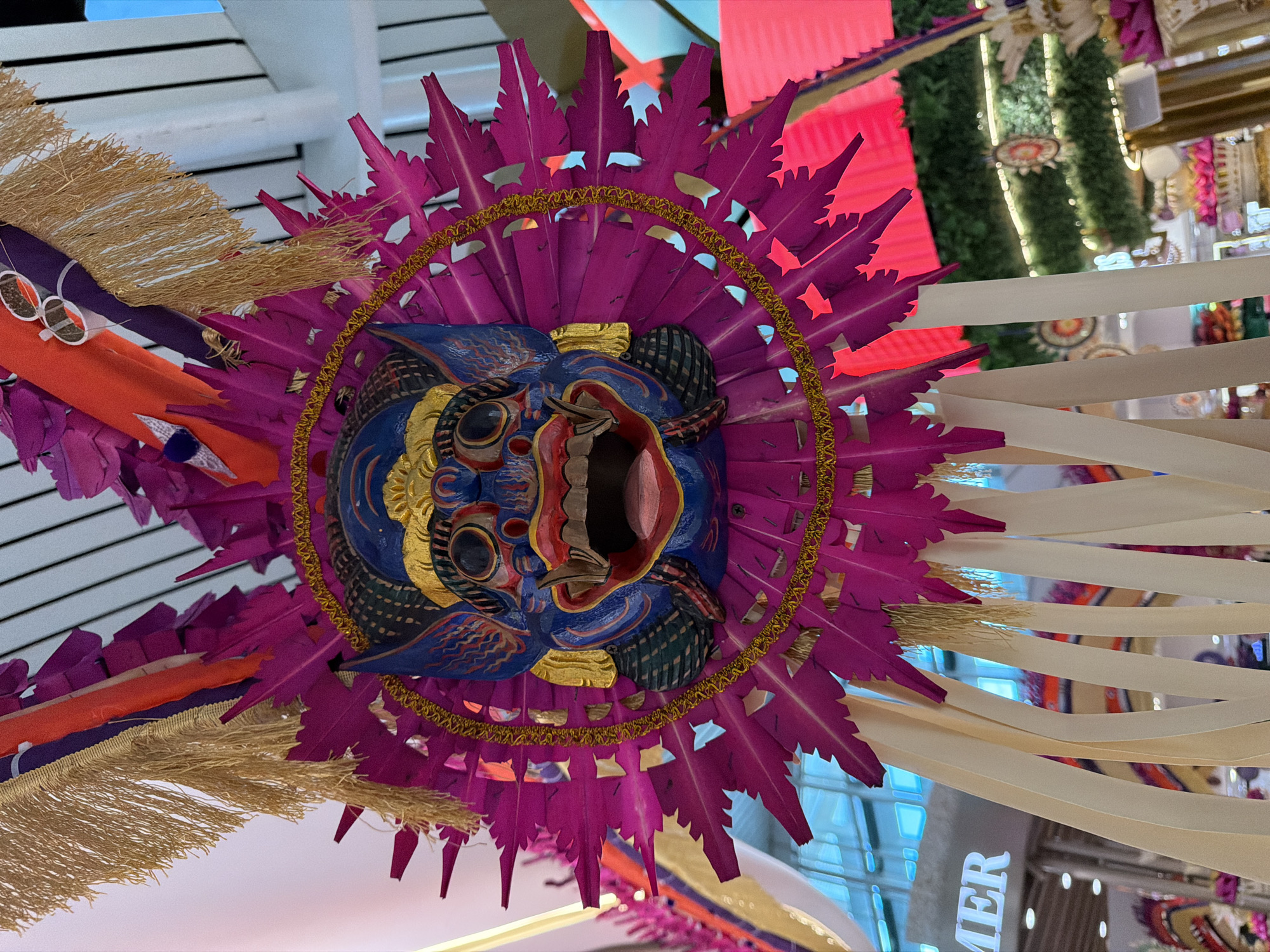 This media file is a close-up, low-angle shot of a vibrantly colored, elaborate mask decoration hanging from a ceiling. The mask itself is the central subject, featuring a fierce expression with a wide-open mouth, sharp teeth, and bulging eyes. It is predominantly blue, with red and pink accents around the mouth and eyes, and gold detailing. The mask is surrounded by a large, fan-like headdress made of numerous pointed, magenta-colored feather-like elements. A thin, gold, braided cord encircles the base of the headdress. Golden fringe hangs from the sides of the headdress, and other decorative elements, including orange and purple fabric strips, are also visible. The background is a blurred mix of colors and lights, suggesting an indoor environment, possibly a marketplace or a decorative display area, with hints of greenery and red lighting. The overall impression is one of festive or ceremonial decoration, likely part of a cultural display. No people are visible in the frame. The setting appears to be an indoor location in Kuta, Indonesia, suggested by the decorative style and context. The lighting suggests it could be daytime or artificially lit indoors. The word "MER" is visible in large, white capital letters in the background, possibly part of a sign or advertisement.