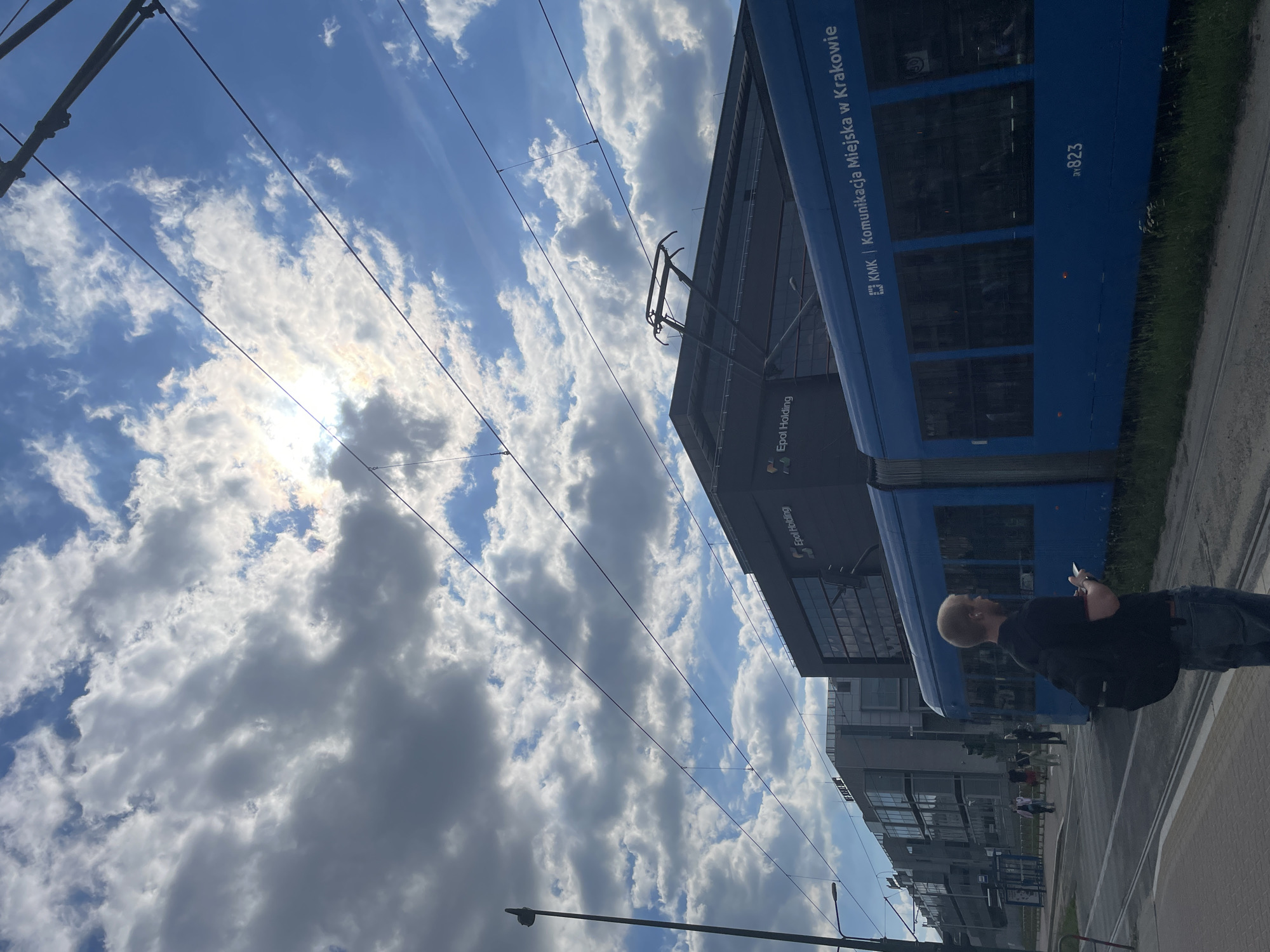 A blue tram runs along a street in Kraków, Poland under a partly cloudy sky. A fair-skinned man with a bald head and beard is on the sidewalk, looking at his phone. Other people are walking behind him. The tram has "KMK | Komunikacja Miejska w Krakowie" written on its side, along with the number 823. In the background are buildings with glass windows. Overhead, electric wires for the tram extend across the sky.