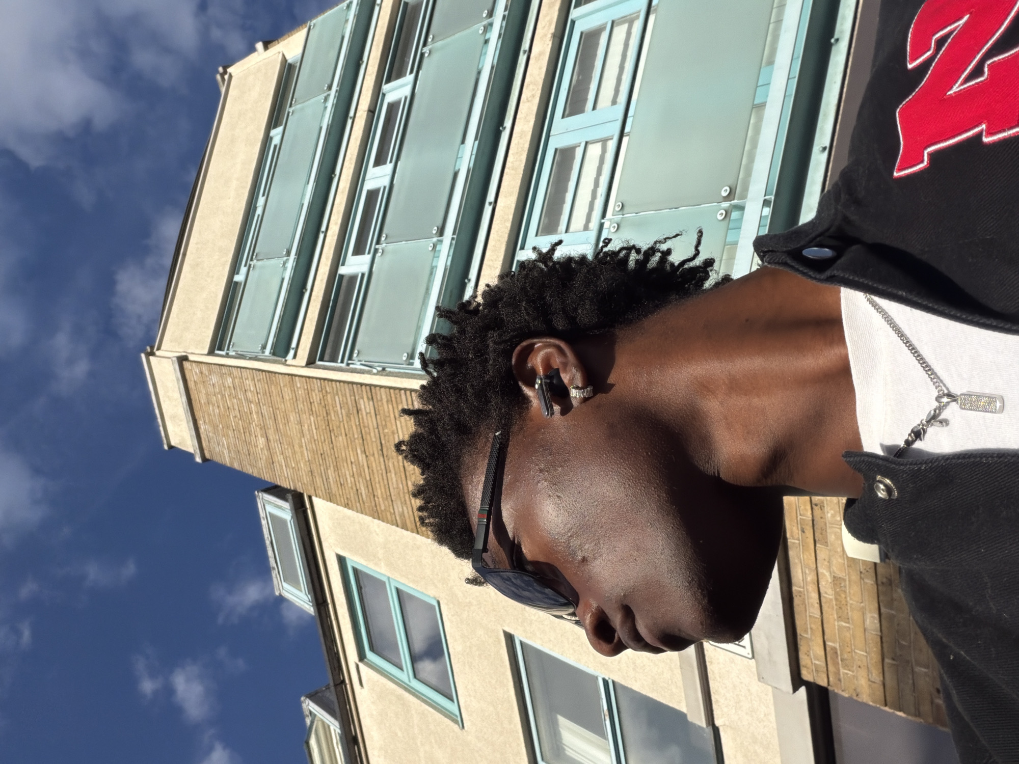 A low-angle portrait of a Black man with dreadlocks wearing sunglasses, a white t-shirt, a silver chain, and a black jacket with a red and white graphic on the upper right. He is wearing a silver earring and an earpiece in his left ear.
He stands in front of a building with multiple floors and pale yellow brick. Windows with blue frames and mirrored or translucent blue panels cover most of the building's facade. The sky is visible in the upper left corner, with some light clouds. The photo appears to have been taken during the day, with ample sunlight. Based on the architecture, the location could be in London, United Kingdom.