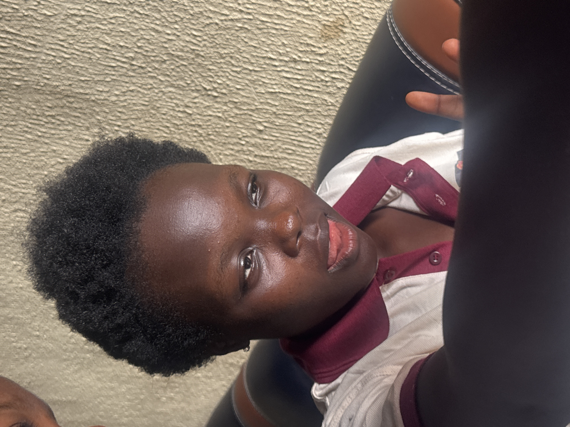 A dark-skinned individual with short, dark hair is photographed indoors against a textured beige wall. The person is seated, possibly in a chair with a brown and black seat, and is wearing a white shirt with a dark red collar and button placket. An arm, partly obscuring the view, is also present in the foreground. The location in Onitsha, Nigeria, cannot be determined from the provided image.