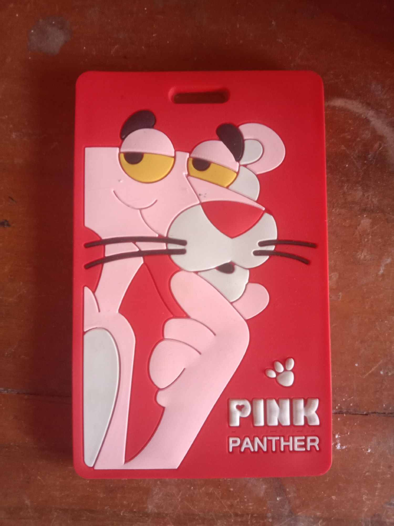 A red identification card featuring a raised relief of the Pink Panther character is lying on a wooden surface. The Pink Panther is depicted from the waist up, with its head turned slightly to the left. Its characteristic pink body is outlined in white, and its eyes are yellow with black pupils. The words "PINK" and "PANTHER" are embossed below the character in white lettering, with a small paw print above "PINK." The wooden surface appears to be a table or desk, with visible grain and some minor surface imperfections. The background is out of focus but suggests an indoor environment. The image is an overhead shot of the object.