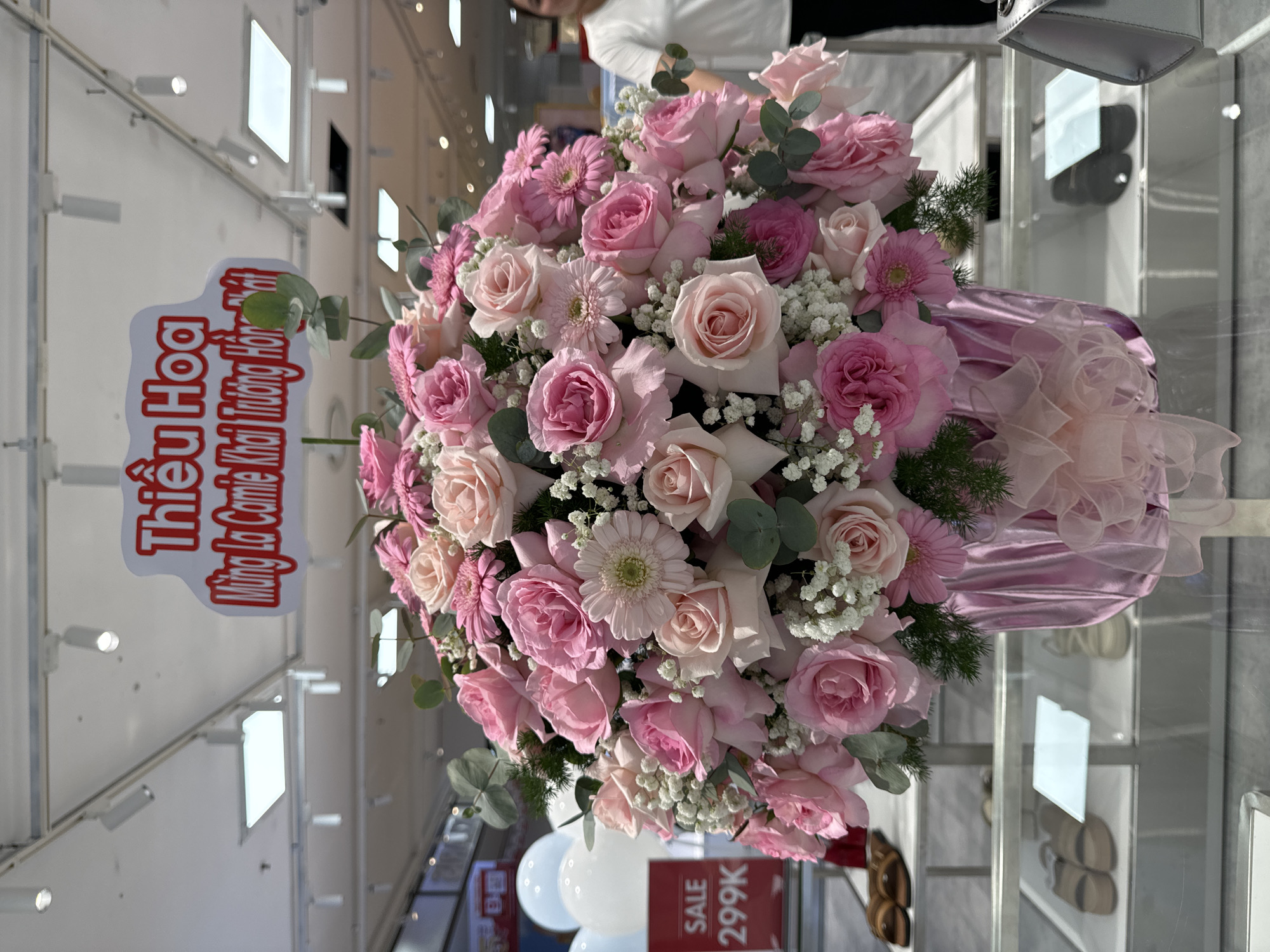 A vibrant floral arrangement, predominantly pink and light pink roses and gerbera daisies, is the central focus of the image. The flowers are densely packed and arranged in a large, light pink vase adorned with a satin ribbon bow. A white sign with red lettering is positioned above the bouquet, reading "Thiếu Hoa Mừng Lễ Khai Trương Hồng Phát" in Vietnamese, indicating a congratulatory message for a grand opening. The arrangement is placed on a glass shelf in what appears to be a retail setting, possibly a store or a mall. In the background, out-of-focus elements suggest a commercial environment with lighting fixtures on the ceiling and shelves displaying merchandise. A red sign with "SALE 299K" is visible to the left of the bouquet, further reinforcing the retail context. A woman in a white shirt is partially visible on the right side of the image, looking towards the left. The overall scene suggests a celebratory event or a store display in Phu Thuy Ward, Vietnam.