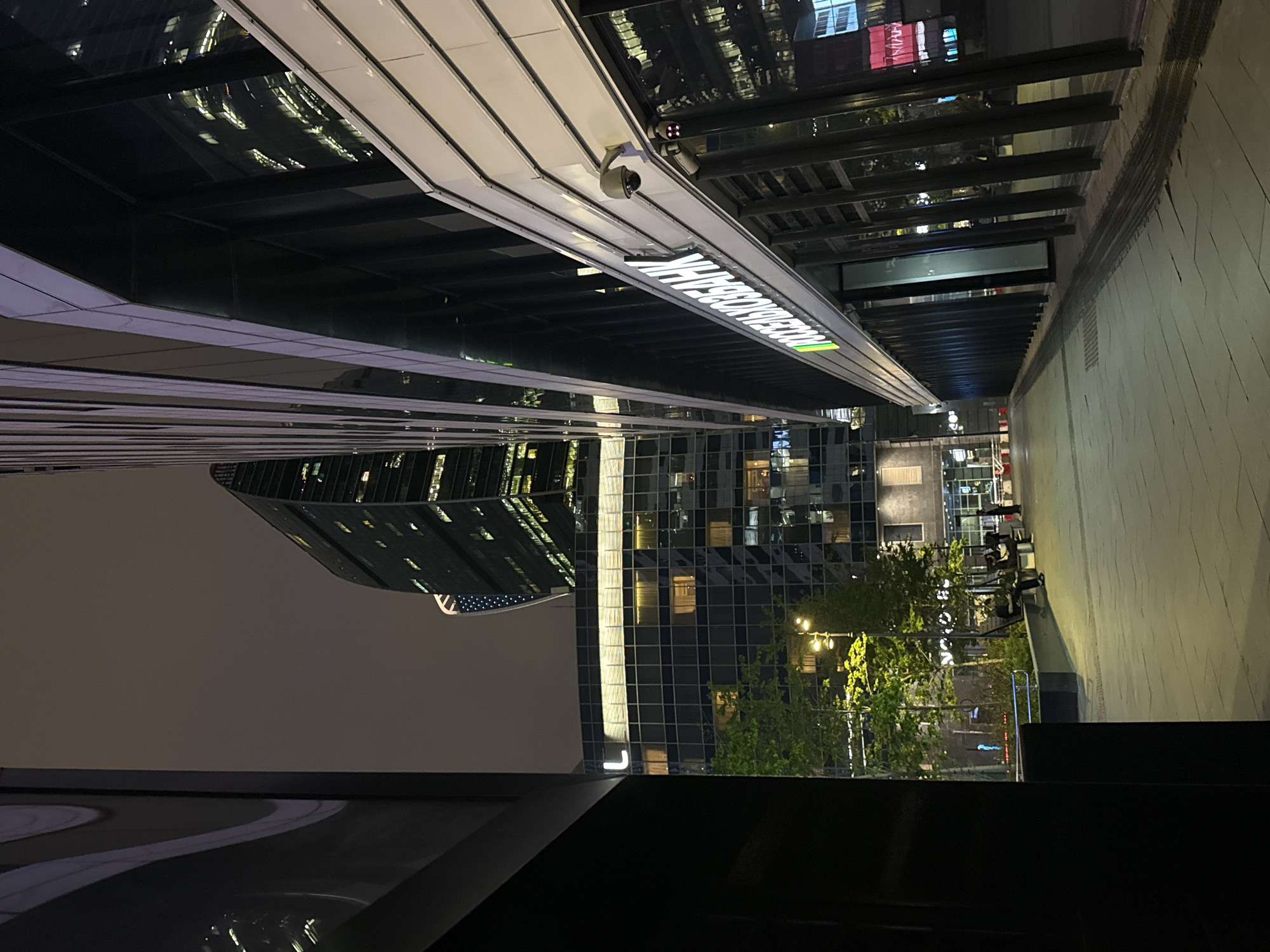 At night in Khimki, Russia, the image captures a cityscape. A tall building with reflective windows dominates the view. The building's architecture is modern, and it seems to reflect the surrounding lights. The night sky is dark, with low light conditions. The image also shows the street and some passersby. A few trees are visible as well. A close-up reveals the architectural details of the building.