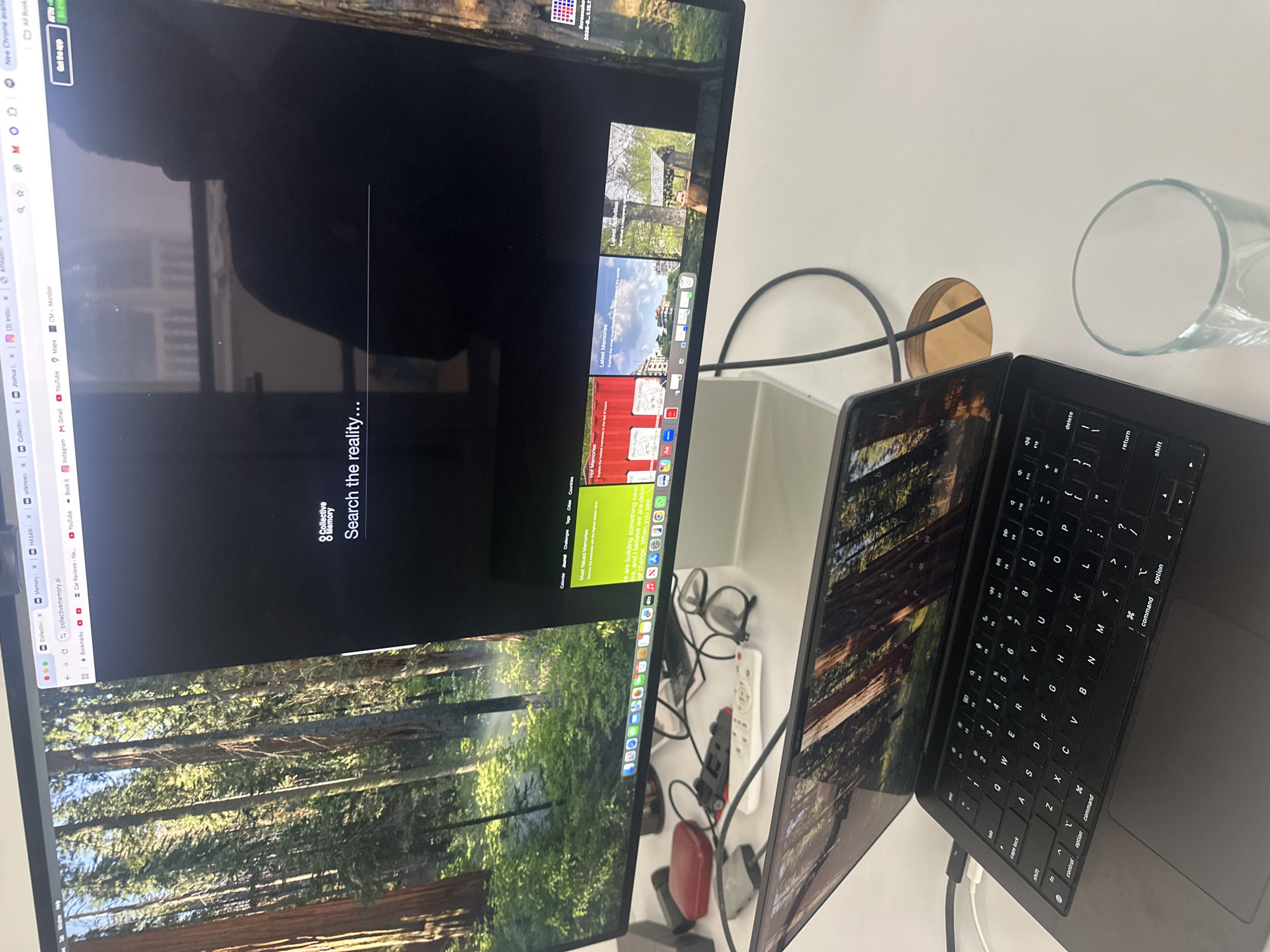 The image shows a white desk with two laptops, a glass, and various small objects. A large monitor dominates the scene, displaying a webpage with the URL "collectivememory.al" and the search prompt "Search the reality...". The background of the webpage features a forest scene. The laptop in the foreground is black and features a standard keyboard layout. Cables are visible connecting the laptop to the monitor and other devices. Scattered on the desk are glasses, a remote control, a toy car, a red object, and other miscellaneous items. The scene appears to be set indoors, likely in an office or workspace. The image does not provide enough detail to determine a specific location in Tel-Aviv, Israel.
