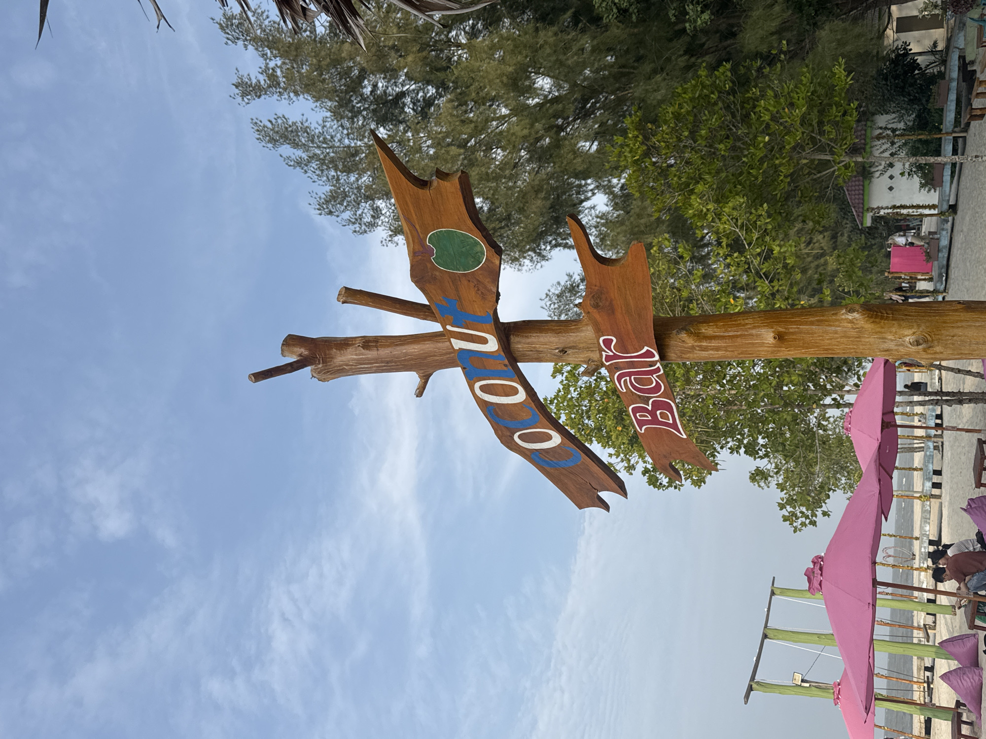 This image depicts a wooden signpost on a beach in Unknown, Indonesia, advertising "COCO" and "Bar." The sign for "COCO" is an arrow shape with the text painted in blue and white, featuring a green circular image of a coconut. The sign for "Bar" is also an arrow shape with the word painted in red and white. The signpost is made of a rough, natural wood trunk. In the background, there are lush green trees, a small white building, and several pink beach umbrellas providing shade. A beach swing set is visible on the left side of the frame. The sky is clear with some scattered clouds. A few people are seated at tables under the umbrellas, and some beanbag chairs are arranged on the sand. The overall scene suggests a relaxed beachside atmosphere.