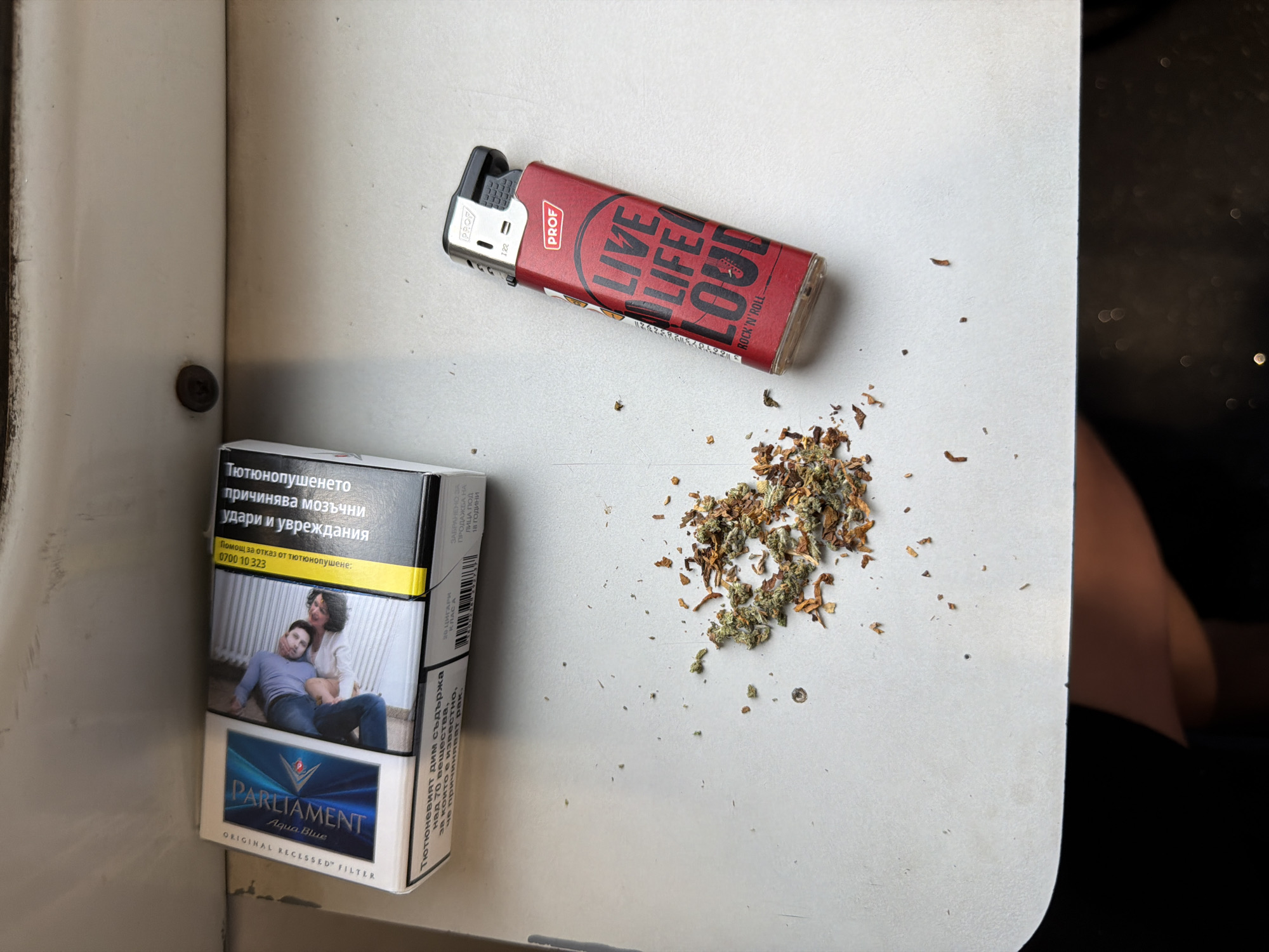 An overhead shot captures a pack of Parliament Aqua Blue cigarettes, a red "PROF" brand lighter, and a small pile of mixed tobacco and dry green plant material on a light-colored surface. The cigarette pack, positioned in the lower-left corner, features a graphic health warning depicting a man reclining on a woman, accompanied by Bulgarian text.
The text on the cigarette pack reads: "Тютюнопушенето причинява мозъчни удари и увреждания" (Smoking causes strokes and damages), "Помощ за отказ от тютюнопушене: 0700 10 323" (Help to quit smoking: 0700 10 323). The brand name "PARLIAMENT Aqua Blue" and "ORIGINAL RECESSED® FILTER" are visible at the bottom. On the side of the pack, additional warnings state: "Тютюневият дим съдържа над 70 вещества, за които е известно, че причиняват рак." (Tobacco smoke contains over 70 substances, known to cause cancer.), "20 ЦИГАРИ КЛАС А" (20 CIGARETTES CLASS A), and "ЗАБРАНЕНО ЗА ПРОДАЖБА НА ЛИЦА ПОД 18 ГОДИНИ" (FORBIDDEN FOR SALE TO PERSONS UNDER 18 YEARS OLD).
The red lighter, angled in the upper-middle section, displays "PROF" on its side and prominently features the phrase "LIVE LIFE LOUD" with "ROCK 'N' ROLL" underneath. A partial number "122" is also visible on the lighter's metal casing. The dry plant material, a mixture of brown tobacco shreds and green flakes (possibly cannabis), is clustered to the right of the cigarette pack and below the lighter. The setting appears to be indoors, possibly on a table or shelf, with a dark, blurred background element on the right side of the frame. A screw head is visible embedded in the surface on the left.
