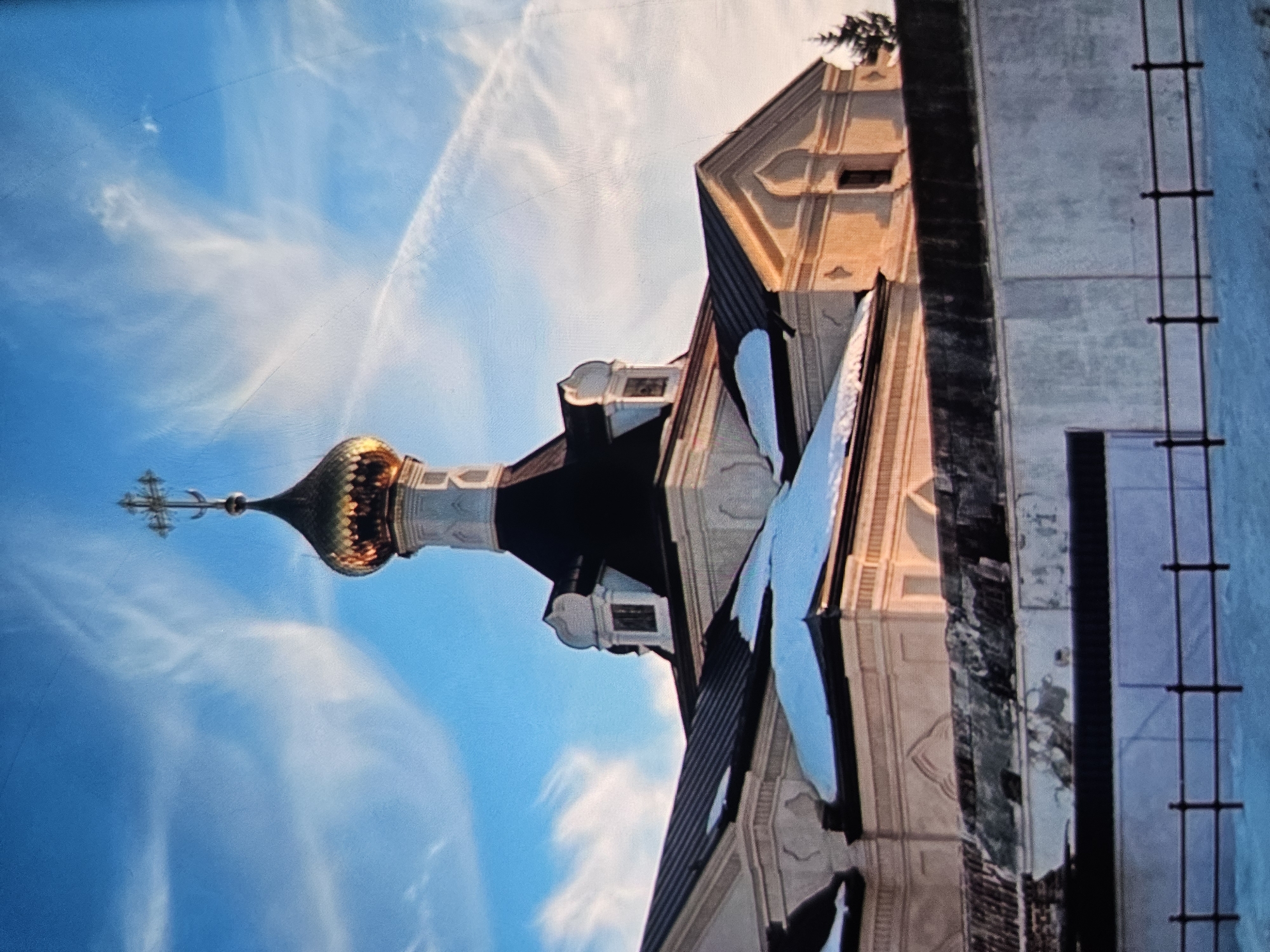 The image shows the gilded dome of a church, topped with a cross, against a bright blue sky with wispy white clouds. The church has dark sloped roofs, some of which are partially covered with snow. A section of a light-colored building with arched windows is visible to the right. In the foreground, a stone wall or fence runs across the scene, with a dark railing in front of it. The ground is covered in snow. A solitary evergreen tree stands to the right of the church. The architecture suggests a Russian Orthodox church. This scene is located in the city of Vladimir, Russia.