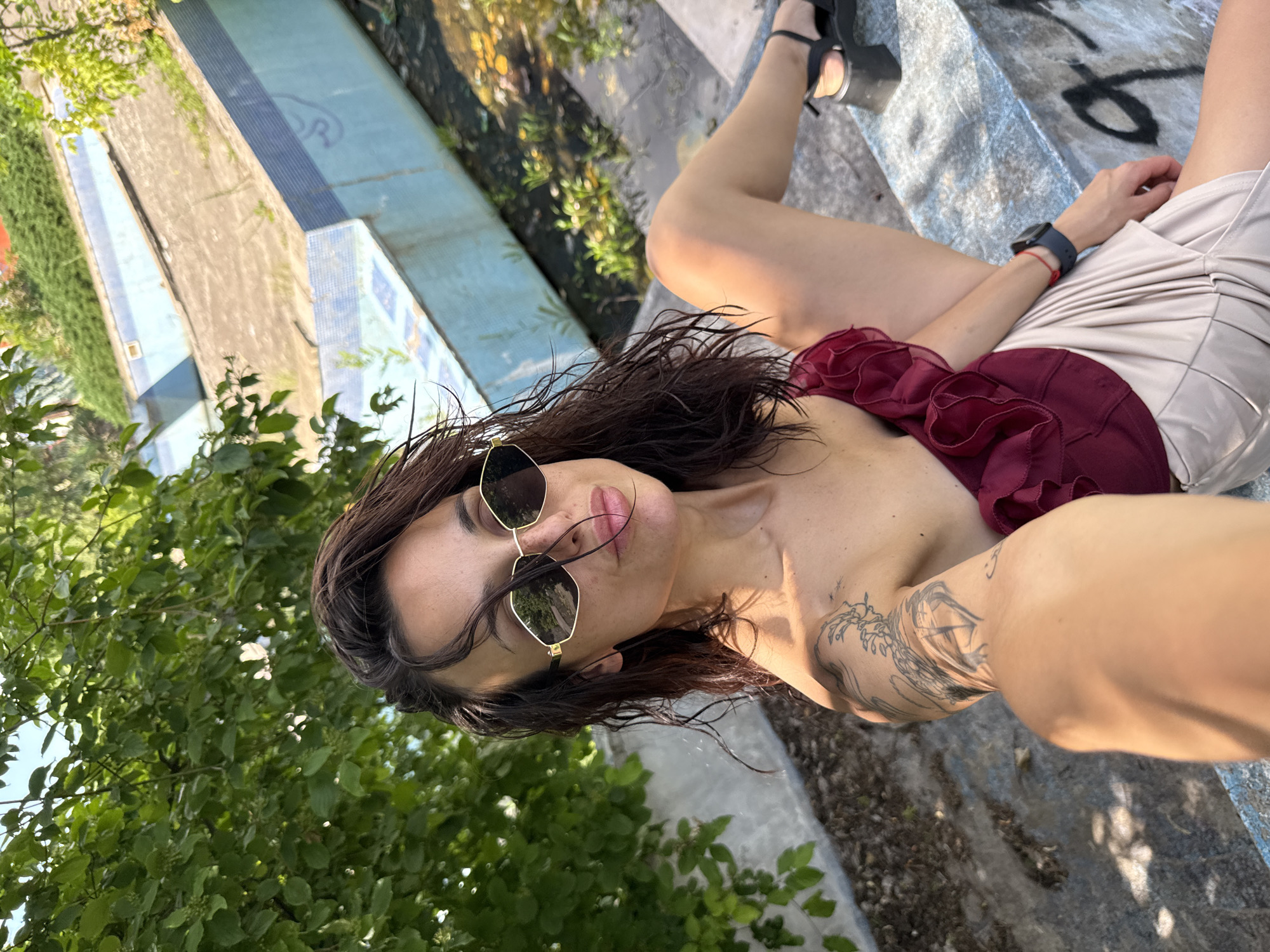 A woman with long, dark, wet hair and sunglasses is posing for a selfie in a disused outdoor swimming pool area in Pleven, Bulgaria. She is wearing a burgundy ruffled top and a light-colored skirt. Her left arm has a tattoo of a dragon. The pool area appears to be overgrown with vegetation and shows signs of disrepair, with cracked concrete and some standing water. The sky is visible through the leaves of a tree in the upper left corner.