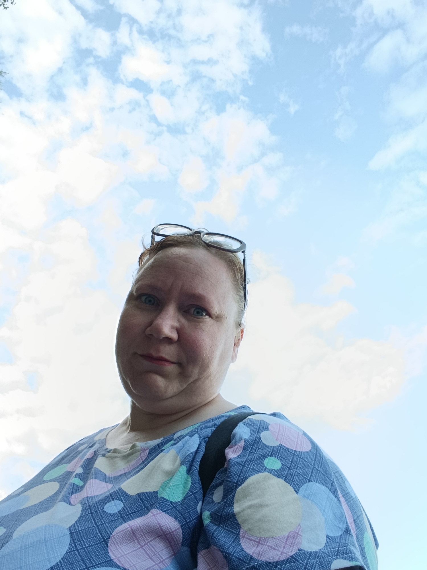 In this low-angle, eye-level shot, a woman with light skin, red hair, and blue eyes is looking at the camera with a slightly quizzical expression. She is wearing glasses perched on top of her head and a blue and white patterned top adorned with colorful circles in shades of pink, mint green, and yellow. The background consists of a bright blue sky filled with fluffy white clouds, suggesting it is daytime with fair weather. The woman appears to be outdoors, possibly in a park or open area, given the expansive sky. The image was taken in Chaikovsky, Russia.