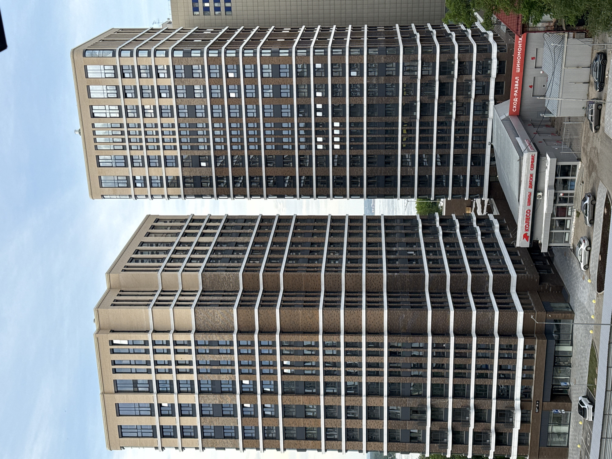 Two tall, modern buildings with a distinctive architectural design are featured prominently. The buildings have a patterned facade consisting of rows of windows and brown brickwork, separated by white vertical lines. These white lines create a slightly "wavy" or stepped appearance on the exterior of the building.
To the right of the buildings, there is a smaller building identified by signs in Russian, indicating it is a car service or tire shop. The sign reads "КОЛЕСО ШИНЫ ДИСКИ СЕРВИС СХОД-РАЗВАЛ ШИНОМОНТАЖ". Several cars are parked around the building.
The sky is visible in the background. The perspective suggests the photo was taken from an elevated position, looking downward at the buildings. The image depicts an urban environment, likely the city of Perm, Russia.