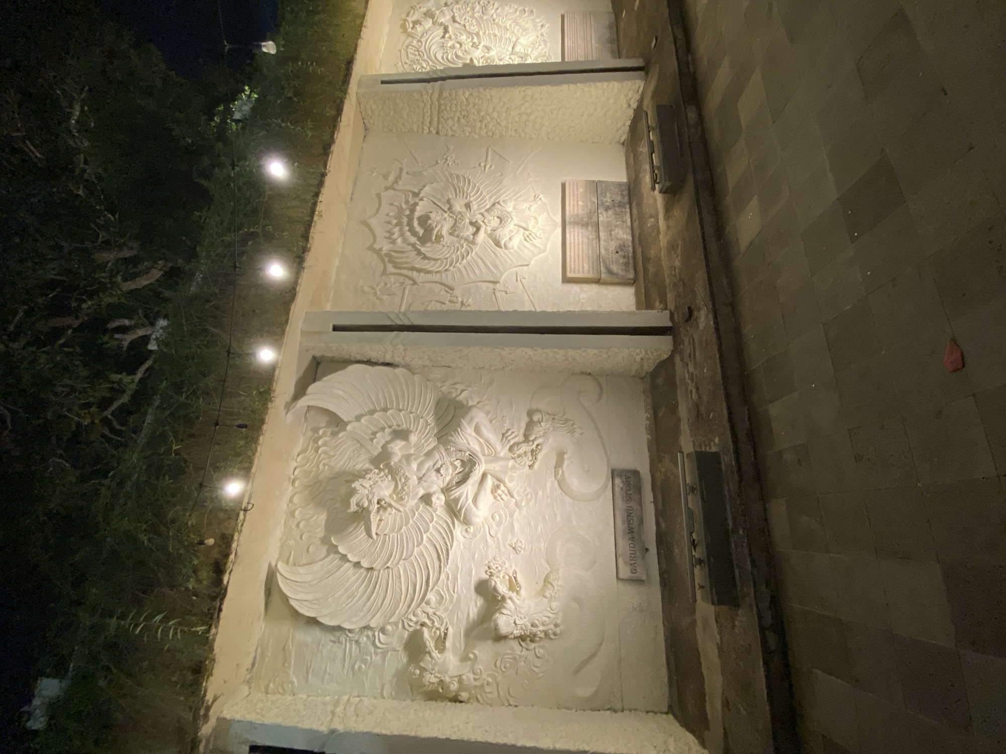 The image depicts a nighttime view of an outdoor relief sculpture display featuring mythological figures. The main subject is a large white relief of Garuda, a mythical bird-like creature, with outstretched wings and a human-like torso. The sculpture is illuminated, casting shadows and highlighting the intricate details of the carving. The relief is mounted on a textured white wall, which is part of a larger structure that appears to be a monument or an exhibition space.
Below the Garuda relief, a rectangular plaque is visible with the text "GARUDA-WISNU STORY" etched onto it. This plaque is placed on a dark, flat surface that runs along the base of the wall. In front of the plaque, there are several dark, rectangular blocks or bases, some of which are partially submerged or appear to be part of the foundation.
The wall is divided into sections by vertical pillars, and to the right of the main Garuda relief, another similar relief is partially visible, suggesting a series of these sculptures. The background behind the wall is a dark, overgrown slope covered in dense foliage, with trees and vines. String lights, each with a visible bulb, are suspended above the wall, providing additional illumination to the scene and casting a warm glow. The foreground consists of a paved area made of dark rectangular tiles, characteristic of a walkway or plaza. A few scattered items, including a piece of red debris, are visible on the pavement.
Based on the inscription "GARUDA-WISNU STORY" and the cultural significance of Garuda in Indonesian mythology, it is highly probable that this scene is located in Bali, Indonesia. Given the mention of Kuta Selatan, this specific location within Bali is a possibility. The overall ambiance is serene and somewhat mystical, enhanced by the nighttime setting and the artistic presentation of the reliefs.