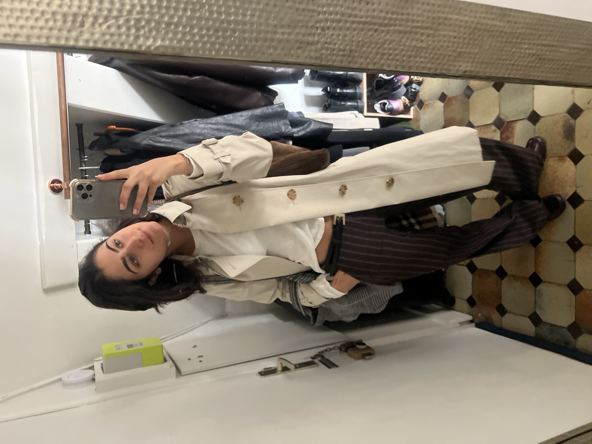 A woman takes a mirror selfie in a room with cream and brown patterned tile flooring. The mirror is framed with a dull gold material. The woman holds a cell phone in her right hand, her right arm partially obscured by a light tan trench coat she is wearing. The coat is open, revealing a white shirt and dark brown pinstripe pants. A black belt is visible on her waist. Her dark brown hair falls over her left shoulder.
Behind the woman and the mirror are clothing items hanging in what appears to be a closet or storage space. Leather jackets and other garments are visible. The counter on which the woman leans holds an electrical outlet, a small white electronic device with a lime green top, and a small padlock on a chain.
Given the architecture and tile, the location could be a residential or retail space, possibly located in Paris, France, due to the architectural style.