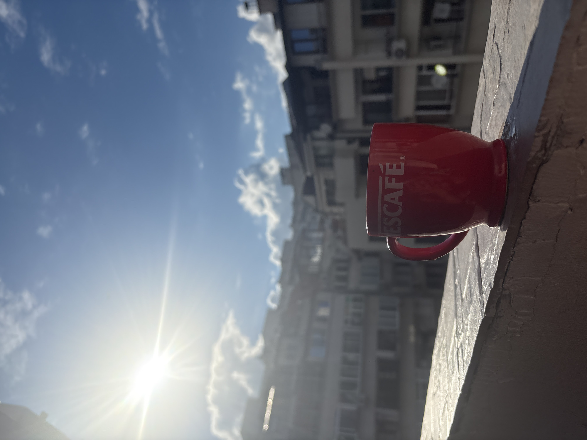 A red Nescafé mug sits on a textured, light-colored surface in the foreground. The mug has the Nescafé logo printed on it in white. Behind the mug, blurred buildings are visible, suggesting an urban environment. The sky is blue with scattered white clouds, and the sun is shining brightly, casting a lens flare in the upper left portion of the image. The scene appears to be outdoors, possibly on a balcony or windowsill, overlooking a cityscape. The location is Burgas, Bulgaria.