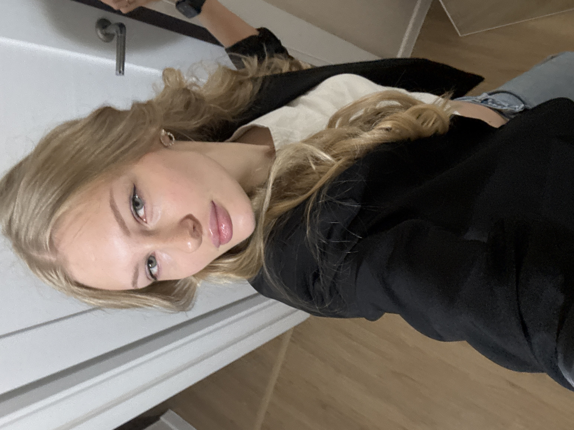 A young woman with blonde hair is taking a selfie in what appears to be a residential hallway, possibly in Perm, Russia. She is wearing a black blazer over a cream-colored top and light-wash jeans. Her left arm is extended to hold the camera, and her right hand is visible near a door handle. The hallway has light-colored walls with vertical paneling and wooden flooring.
