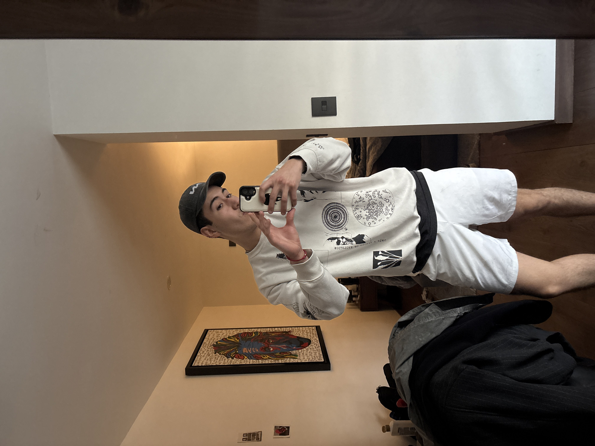 A young male is captured in a mirror selfie, taken indoors in what appears to be a modern home in San Bernardino Tlaxcalancingo, Mexico. The man stands facing the mirror, holding a light-colored smartphone with three rear cameras in both hands, taking his own picture. He wears a grey baseball cap with a white symbol (possibly an 'M' or 'W'), a white long-sleeve sweatshirt with various black graphic designs, and white shorts with a black belt. His lips are pursed, suggesting a playful or "blowing a kiss" expression.
The sweatshirt features intricate black circular patterns with smaller circles inside, abstract shapes, and visible text that reads "EVOLUTION NO ESCUPO A IJO" along with another black abstract graphic. A red bracelet is visible on his right wrist.
The background shows white walls, a dark wooden beam or trim at the top, and light brown wooden flooring. A black light switch is mounted on the wall above the man's shoulder. On a surface to the left, there is a framed piece of artwork depicting a vibrant, stylized head of a fish or mythical creature in red, blue, and yellow tones, with a background that appears to contain illegible text. Below this, two smaller framed items are faintly visible. In the lower right corner, a pile of dark clothing, including a pinstriped jacket, is stacked.