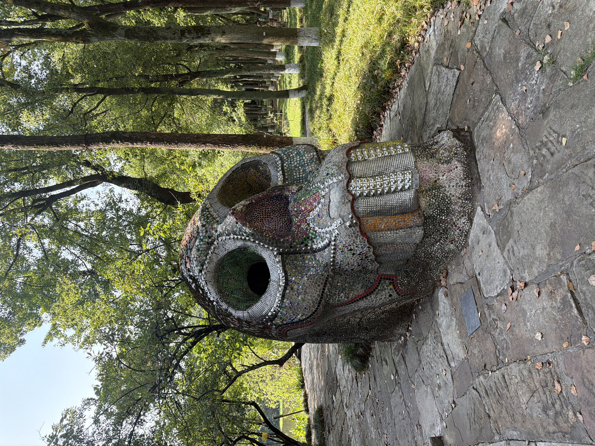 The image shows a large, mosaic-covered sculpture in the shape of a skull, situated on a stone pathway in an outdoor park setting. The skull sculpture is predominantly gray and adorned with various colored mosaic tiles, creating intricate patterns. It has large, hollow eye sockets and a grinning mouth with square teeth. The pathway is paved with irregular stone slabs and is partially covered with fallen leaves.
To the left of the sculpture, a body of water is visible, bordered by lush green vegetation and trees. Behind the sculpture, a dense arrangement of trees forms a backdrop. Some of the tree trunks are painted white at their bases, a common practice in China for pest control and to mark them. The overall environment appears to be a well-maintained park with sunlight filtering through the trees, casting dappled shadows on the ground. The scene is peaceful and suggests a natural or artistic landscape.
This sculpture is located in the Chongqing High-tech Zone, China, specifically in the area of the "City of Stars" cultural and creative industrial park.