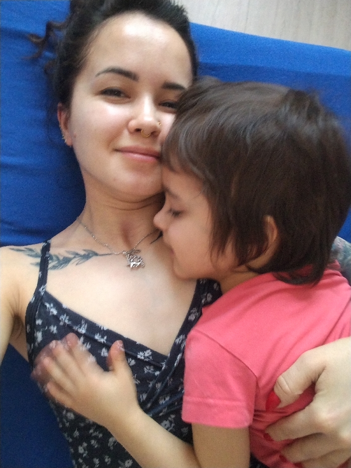 A fair-skinned woman and child lie close together on blue fabric. The woman wears a floral-patterned, spaghetti-strap top and a necklace with an animal pendant. She has dark hair pulled up, a nose ring, and a tattoo on her shoulder. The child, wearing a short-sleeved pink shirt, snuggles against the woman, with a hand placed on her chest. The woman smiles gently, gazing at the camera, while the child's eyes are closed.