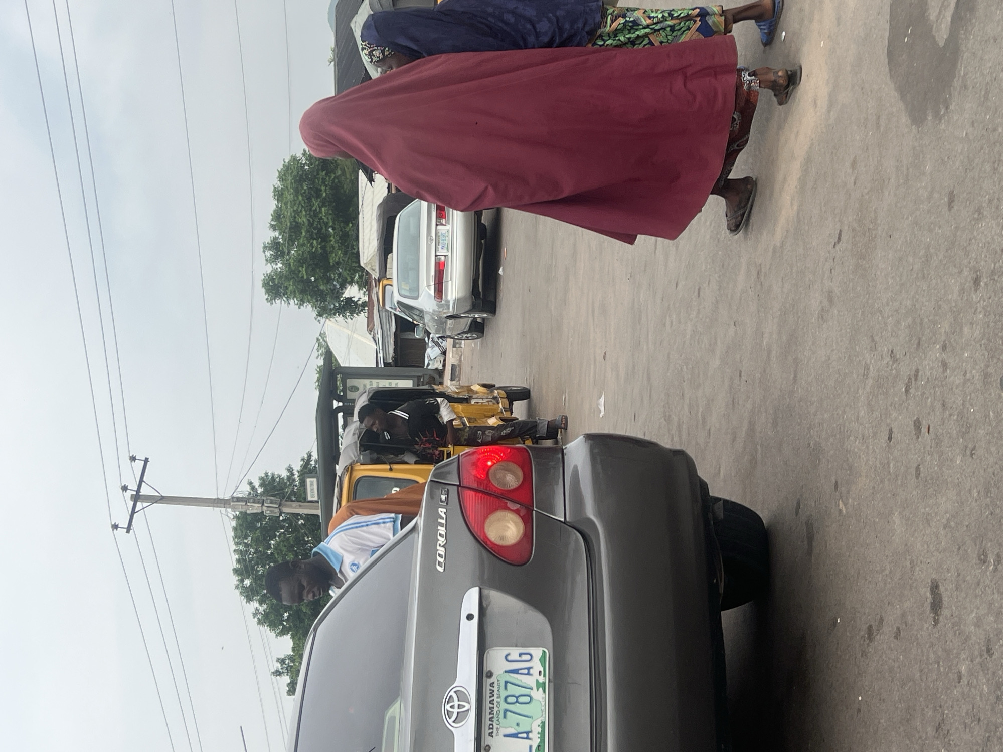 The image depicts a street scene in Howayi, Nigeria. A gray Toyota Corolla, with license plate A-787 AG, is in the foreground. A yellow tricycle and a silver car are positioned behind the Corolla. Two women are walking on the right side of the frame, one in a long red robe and the other in a blue outfit with a patterned skirt. A man is visible in the Corolla. Power lines and trees are in the background. The overall environment appears to be urban.