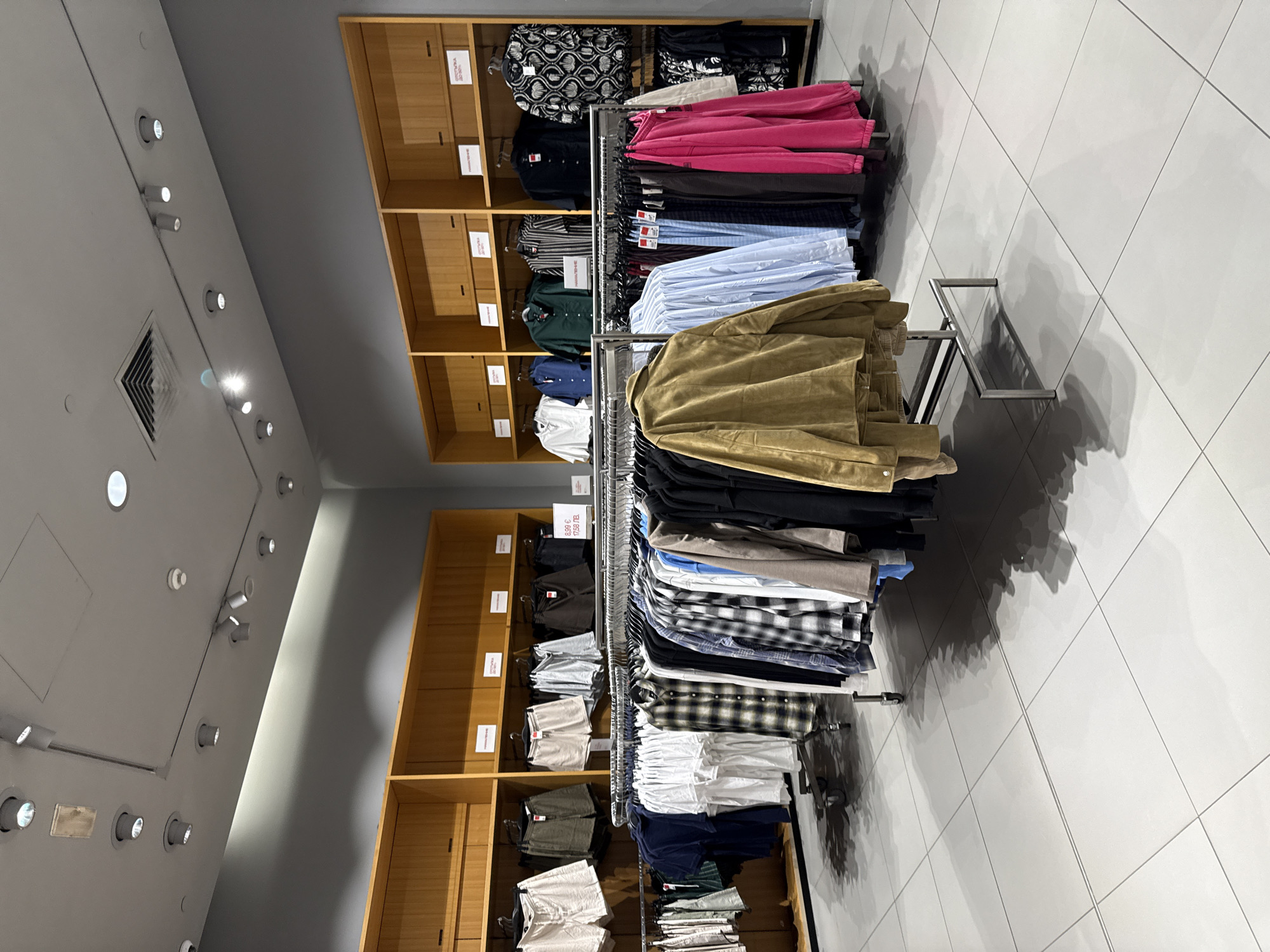 The image shows the interior of a clothing store, likely a retail chain, located in Stara Zagora, Bulgaria. The store features wooden shelves displaying folded garments and racks of hanging clothing, primarily tops and bottoms. The lighting is provided by overhead track lights. The floor is tiled in a light gray color. There are no people visible in the image. The overall scene depicts a well-organized retail environment for apparel.
