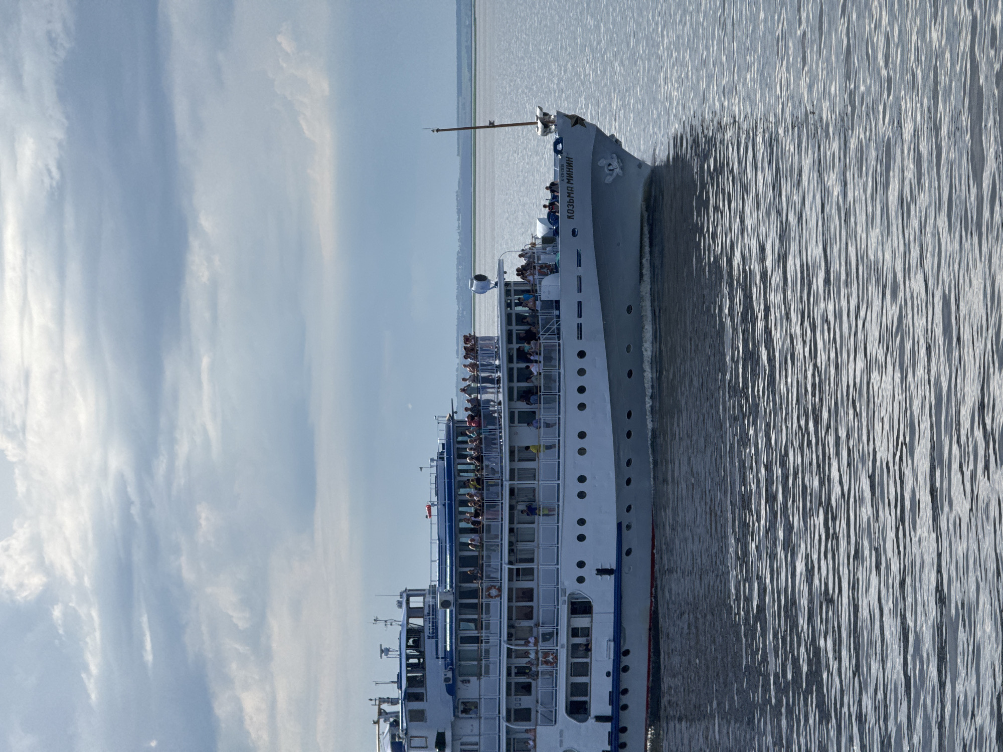 A wide shot captures a multi-decked white passenger ship, the "KOЗЬМА МИНИН," cruising on a large body of water under a cloudy sky. The ship is positioned in the foreground, angled slightly to the right, with its port side visible. Numerous passengers can be seen on the open decks of the ship, some standing and looking out, others engaged in conversation. The ship has several levels of windows and railings, and on the bow, the name "KOЗЬМА МИНИН" is clearly marked in Cyrillic. The water is calm with noticeable ripples reflecting the light. In the distance, a faint shoreline with greenery is visible. The location is identified as Izhevka, Russia, suggesting this is a scene from a river or large lake within or near the city.