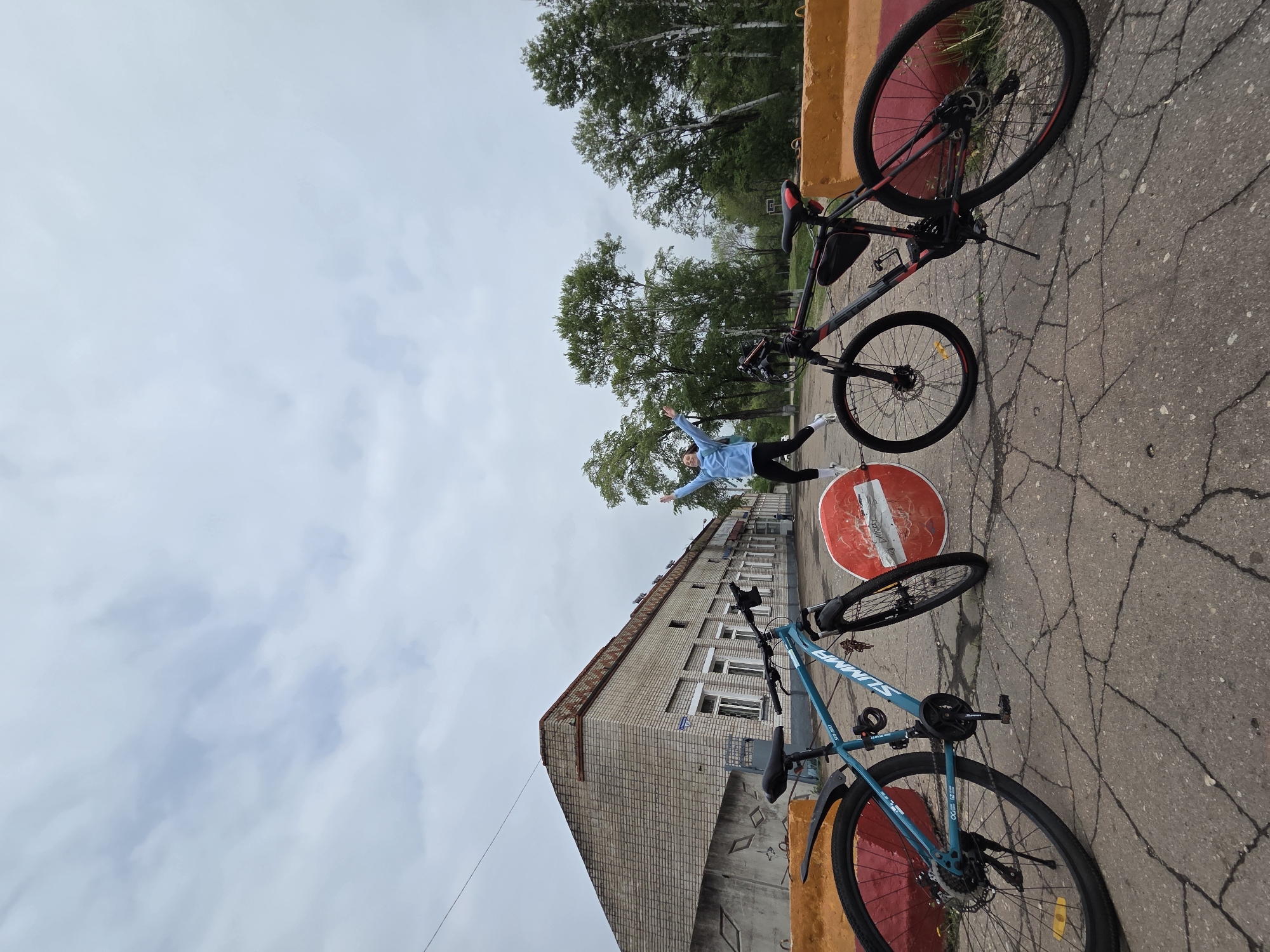 The image depicts an outdoor scene in Birobidzhan, Russia, featuring two bicycles, a person, a building, trees, and a road.
A young adult stands with arms outstretched between the two bicycles. The person wears a light blue jacket, dark pants, and white socks. One bicycle is dark with red accents, and the other is teal. Both are resting against a low, painted barrier, orange and red.
Behind the person and the bicycles is a rectangular, multi-story brick building. The building has several windows. Overhead are trees, and the sky is overcast. The ground is paved and cracked. A round road sign with a white horizontal bar within a red circle is seen at ground level.