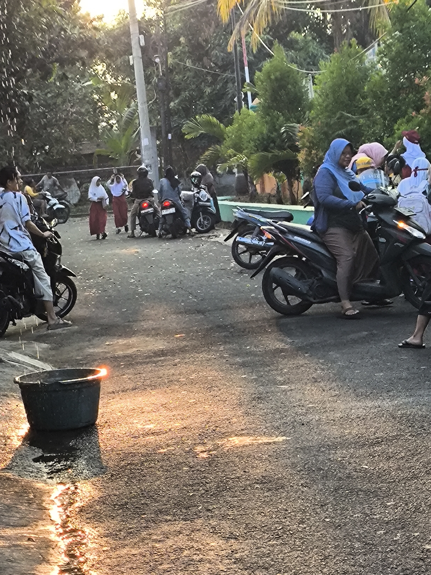 A group of people, some wearing traditional Islamic attire, are gathered on a street in Cilegon, Indonesia. Several individuals are on or near motorcycles. In the foreground, a black bucket is positioned on the asphalt, and light reflects off a wet surface near it. The background shows trees, a utility pole, and buildings, indicating an urban or semi-urban environment. The scene appears to be set during the late afternoon or early evening, as evidenced by the warm, low-angle sunlight.