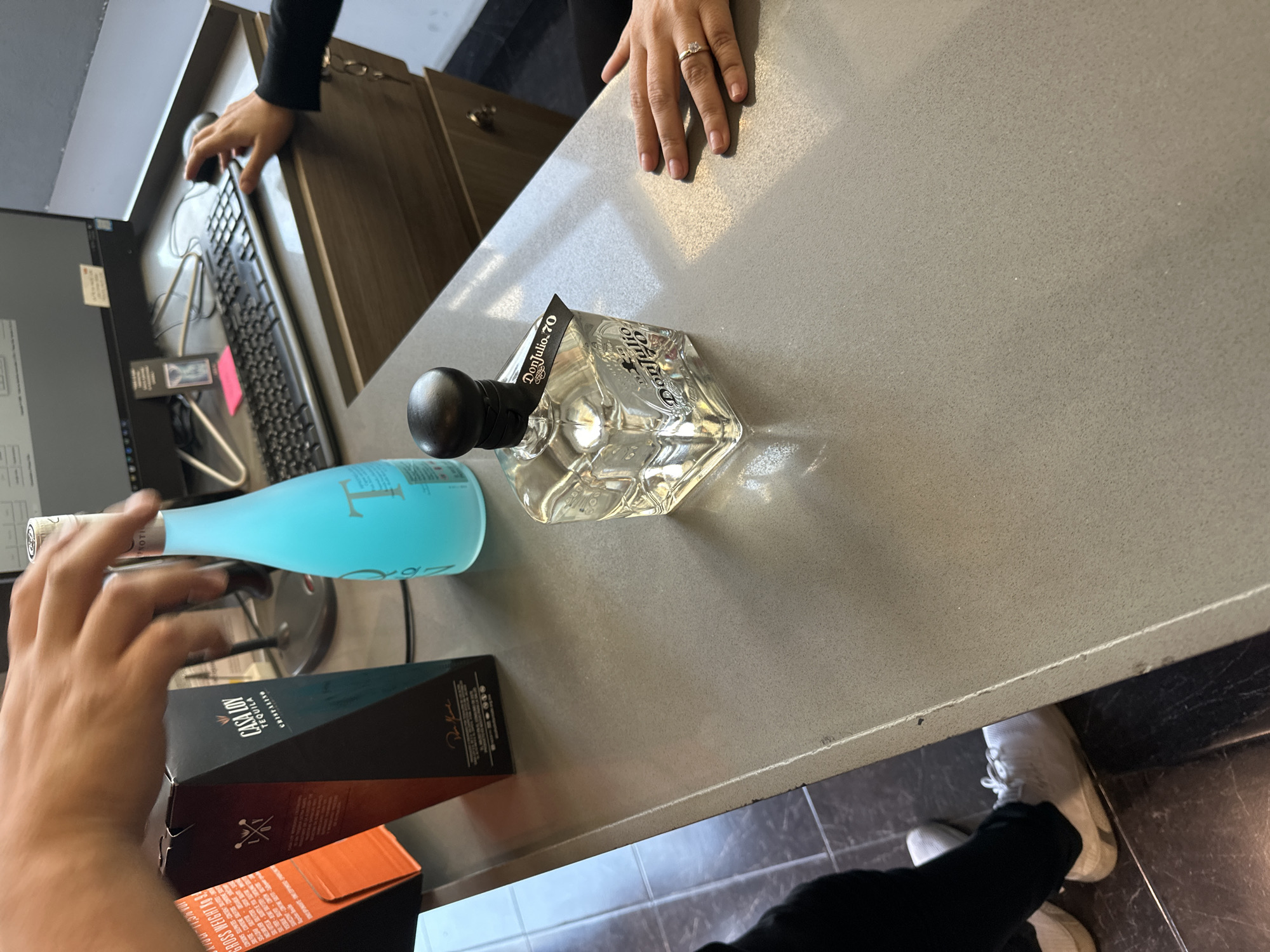 The scene depicts an indoor commercial transaction at a counter, likely within a retail establishment selling liquor. The precise location within Puebla, Mexico, cannot be determined from the visual evidence.
A light gray, speckled countertop extends across the image. On the counter, from left to right, are:
1. A partial view of an orange cardboard box, labeled "GROSS WEIGHT X 5 KG".
2. A dark, faceted cardboard box bearing "CASA LUY TEQUILA" branding.
3. A frosted light blue, slender glass bottle with "TRON" branding partially visible.
4. A clear, square glass bottle of "Don Julio 70" tequila, distinguished by a black spherical stopper and a black neck tag.
Behind these items, a computer workstation is present, featuring a black desktop monitor displaying a software interface, a black keyboard, and a dark gray computer mouse. A small pink sticky note is affixed to the monitor frame.
Two individuals are partially visible:
1. **Individual 1 (Left):** Only the hands, lower legs, and feet are shown. The hands are positioned above the blue bottle and the "CASA LUY" box, in an active posture. This individual wears black pants and white athletic shoes, standing on a dark tiled floor.
2. **Individual 2 (Right):** Only the right hand and forearm are visible. The hand rests on the countertop and operates the computer mouse. This individual wears a dark long-sleeved garment and a ring with a clear stone on the right ring finger.
The background behind the counter includes a dark wood-grain cabinet or desk unit, from which a set of keys hangs on the right side. The wall above the cabinet is light-colored. The lighting appears to be artificial overhead illumination.