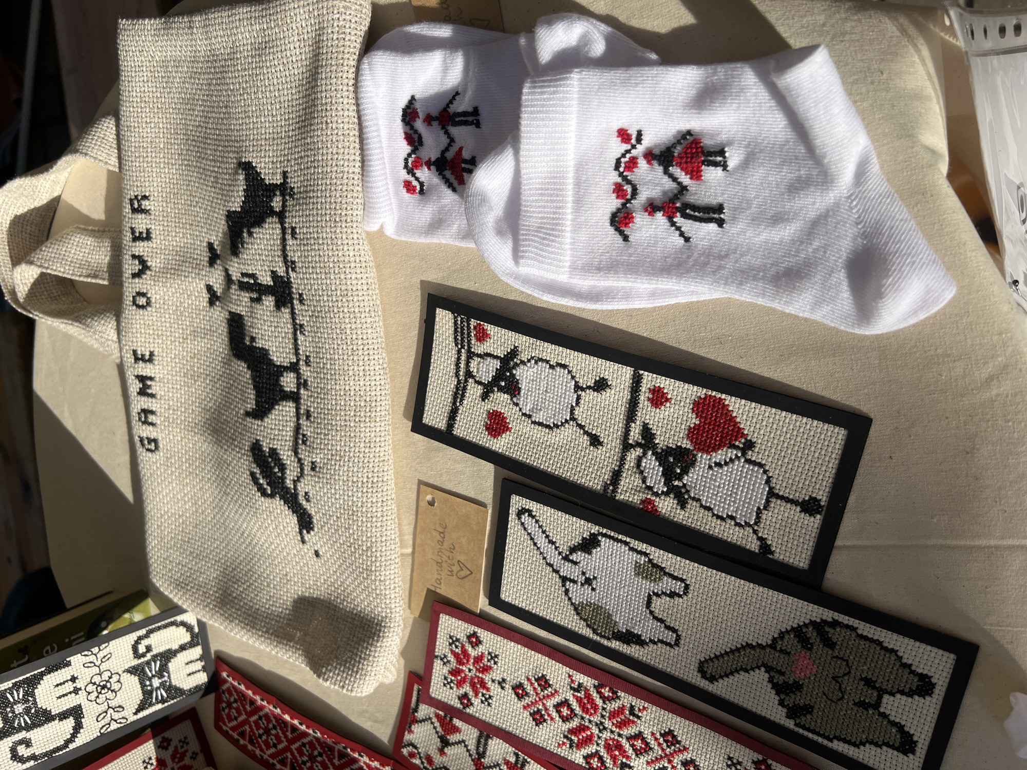 This is a medium-shot, eye-level photograph of handmade cross-stitch items displayed on a light-colored, textured fabric surface, likely a tablecloth or stall covering. The items are arranged to showcase their designs and craftsmanship.
In the background, a canvas tote bag with a raw, natural fiber texture is visible. Embroidered in black thread on the bag is the phrase "GAME OVER" in block capital letters, accompanied by a pixelated black silhouette of two figures, possibly dinosaurs or stylized birds, standing on a line with a cactus in between them.
To the left of the tote bag, a portion of what appears to be a book or another fabric item with a red and white geometric cross-stitch pattern is present. Adjacent to this are several cross-stitched bookmarks. One bookmark features a red and white folk-art-inspired floral or snowflake motif. Another bookmark displays the rear end of a tabby cat with its tail raised. Further to the right, a bookmark depicts a white sheep holding a red heart, and below it, another sheep, also holding a red heart.
In the foreground, two white socks are visible, each embroidered with a pair of stick figures holding hands, with a wavy line of red hearts above them. The right sock has a more detailed design with red accents on the figures' skirts and hair.
A small, light brown tag with rounded corners, appearing to be made of cardstock, is placed between the embroidered bookmarks and the socks. It has the handwritten text "Handmade with ♡" in a cursive font.
The lighting suggests an outdoor setting, with strong sunlight casting shadows across the fabric surface, indicating a bright day. The overall scene suggests a market stall or a craft fair display. There are no people visible in the frame. The location is not specified as being in Chișinău, Moldova.