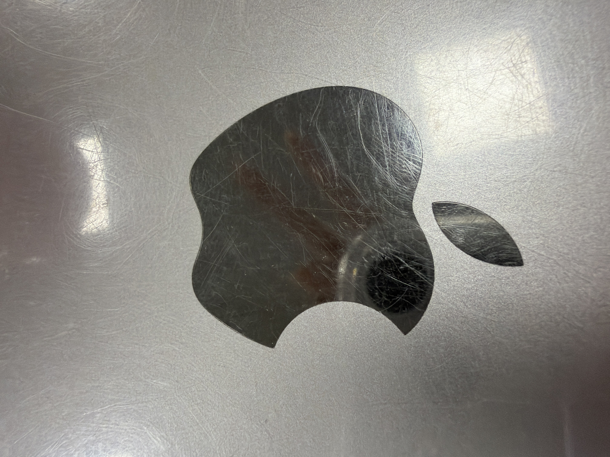 The image depicts a close-up of a silver Apple logo affixed to a surface, likely a laptop or other electronic device. The logo exhibits numerous scratches. The surface surrounding the logo also appears to be silver and has a textured or grainy finish. The reflection of a dark object or person is visible within the logo, suggesting the presence of someone or something in the immediate vicinity. The environment is difficult to discern due to the extreme close-up view. Location is not discernible.