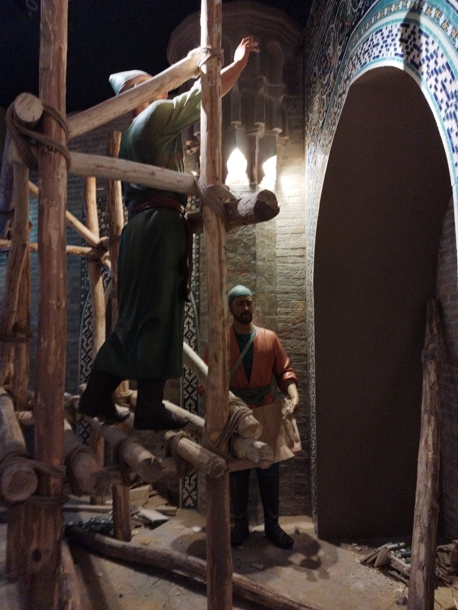 The image depicts a dimly lit indoor scene with two mannequins that appear to be historical figures. One mannequin, dressed in a green robe and a light-colored head covering, stands on a wooden scaffolding structure, reaching upwards with its right hand. The scaffolding is constructed from rough-hewn logs tied together with rope.
To the right, a second mannequin, wearing a brown tunic over a green garment and a blue head covering, is positioned on the ground. This figure holds a piece of fabric or material in its hands and is looking towards the scaffolding. The background shows a brick wall and a large, arched entryway decorated with intricate blue and white tilework. The floor is concrete and appears somewhat dusty, with scattered debris. The lighting is dramatic, casting shadows and highlighting certain elements of the scene. The overall impression is a staged historical diorama.