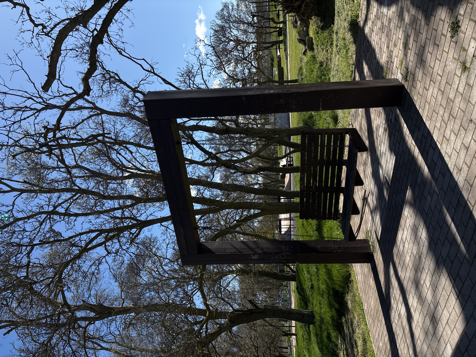 The image shows a wooden bench with an attached dark brown rectangular frame, situated on a brick walkway in a park. Sunlight casts strong shadows from the bench and frame onto the walkway. Behind the bench and frame, there's a view through the frame to trees with bare branches against a blue sky. Green grass is visible on either side of the walkway, and more trees and structures are visible in the background. The location is Chornomorskoe, Ukraine.