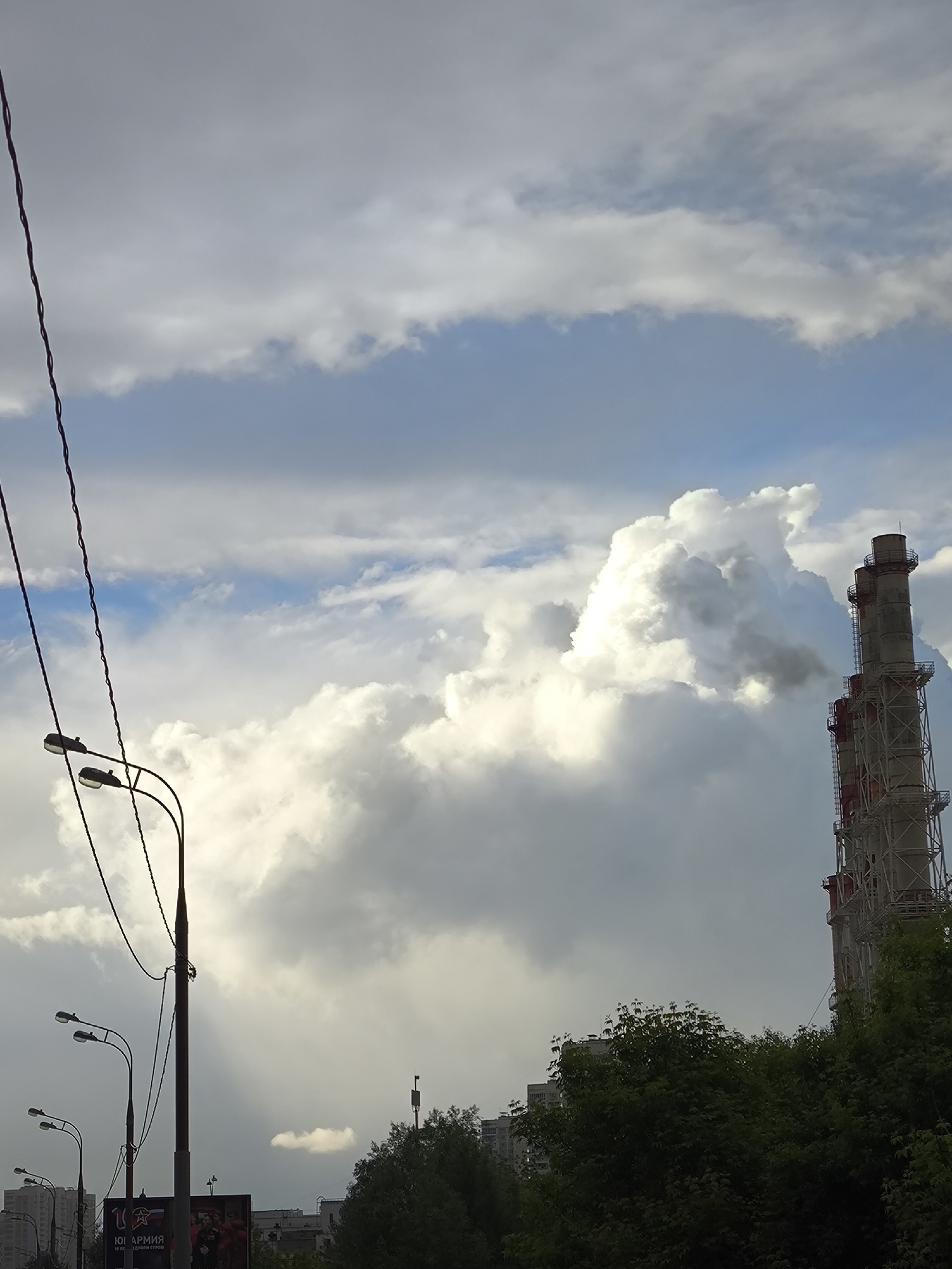 The image depicts an outdoor scene with a mix of natural and industrial elements. In the sky, there are large, billowing white clouds against a backdrop of blue and gray. A tall smokestack structure is present, featuring a red and gray color scheme and a latticework design. Smoke can be seen emanating from the top of the smokestack. The lower portion of the image contains a line of streetlights with curved arms, and power lines are also visible. Green trees fill the foreground and background, suggesting a park or residential area within the city. Buildings can be seen in the distance, partially obscured by the foliage. A billboard is also visible amidst the streetlights.