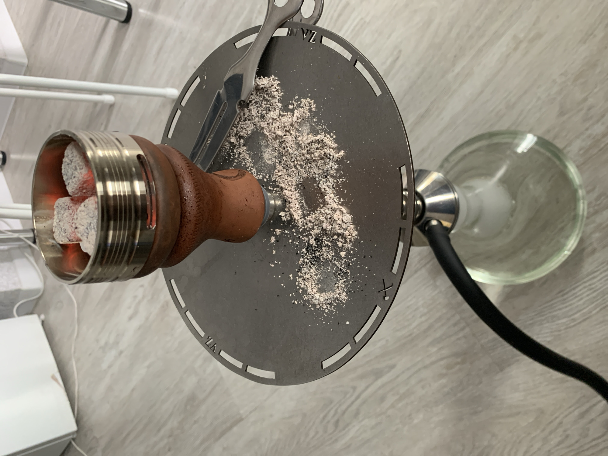 In Otradnoye, Russia, a close-up, eye-level shot captures a hookah setup on a light-colored wooden floor. At the center, a brown ceramic hookah bowl sits atop a metallic tray, supporting a silver heat management device filled with glowing red coals. A small pile of ash and debris is scattered on the tray next to the bowl, with a silver fork resting nearby. A black hose extends from the base of the hookah, which has a clear glass bulb. The background is blurred, showing a white cabinet and white chair legs. The lighting is bright, casting a soft glow on the scene.